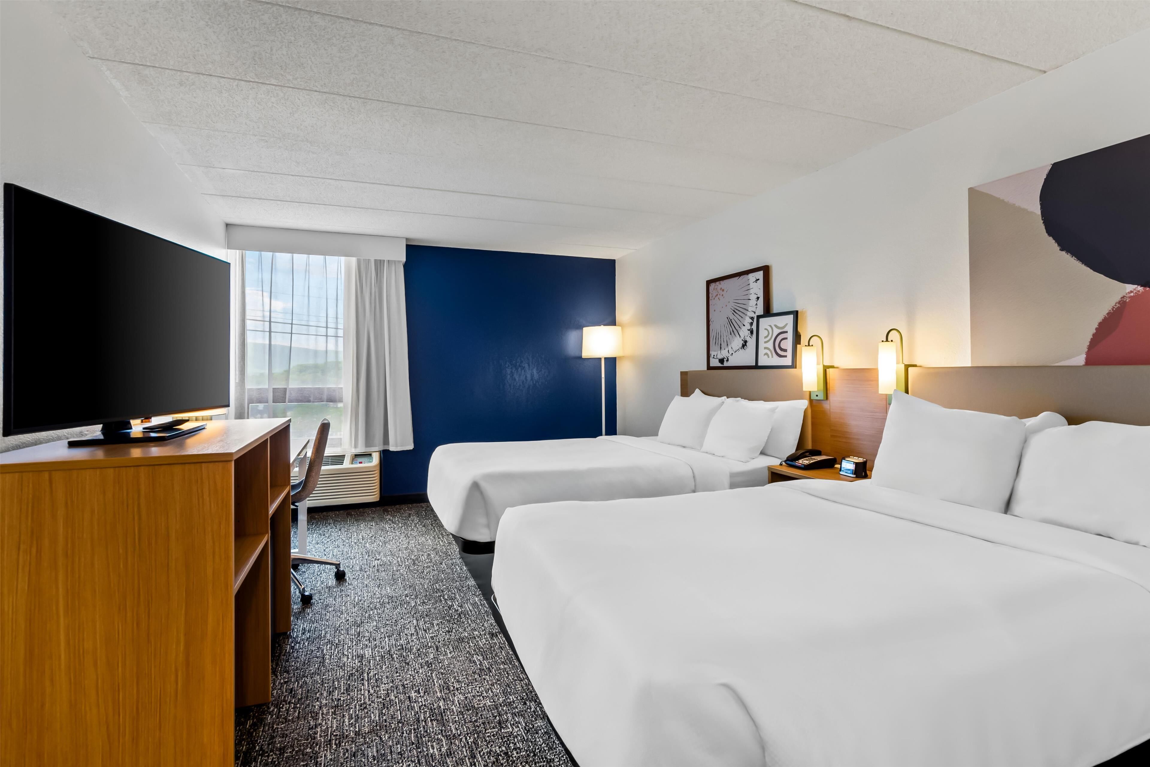 A hotel room with two beds, a desk, a TV, a lamp, and a large window.