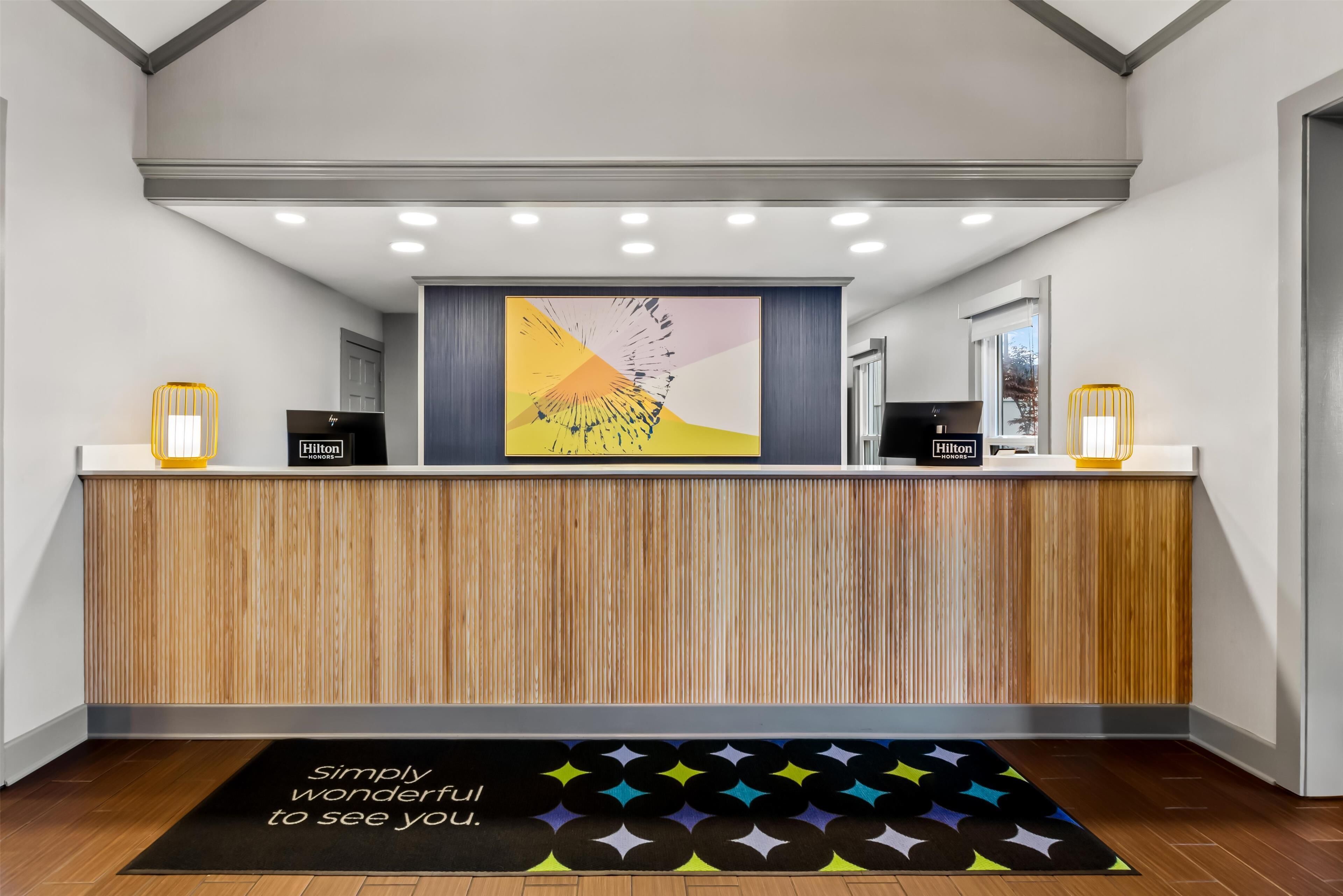 Un mostrador de recepción con diseño de paneles de madera, una obra de arte abstracta y colorida sobre él, y una alfombra negra con una cita motivacional.