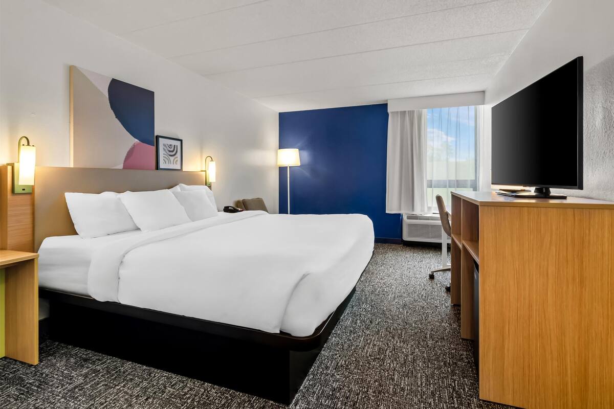 A hotel room with a bed, a desk, a flat-screen TV, and a window with curtains.