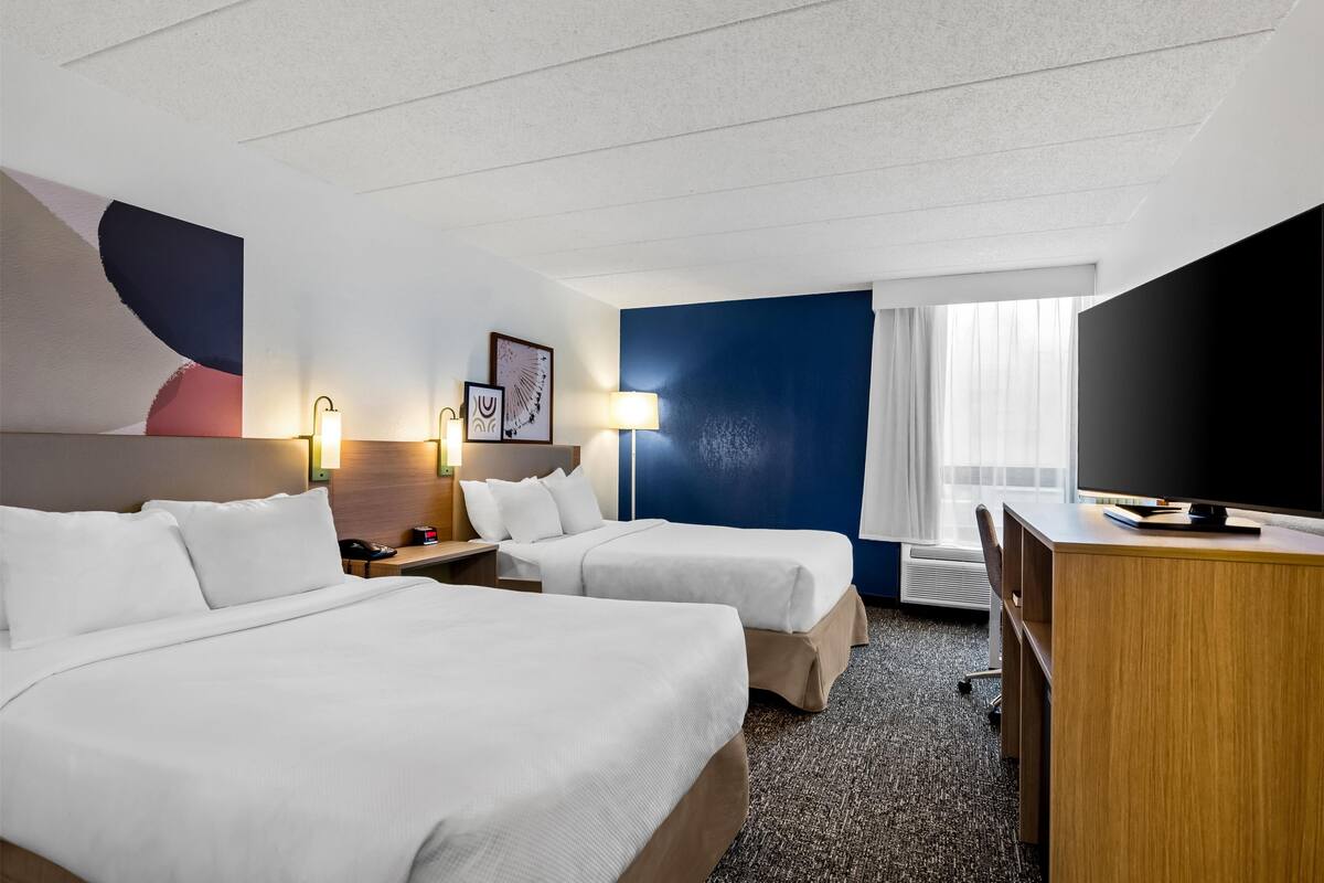 A hotel room with two beds, a desk, a TV, and a large window with curtains.