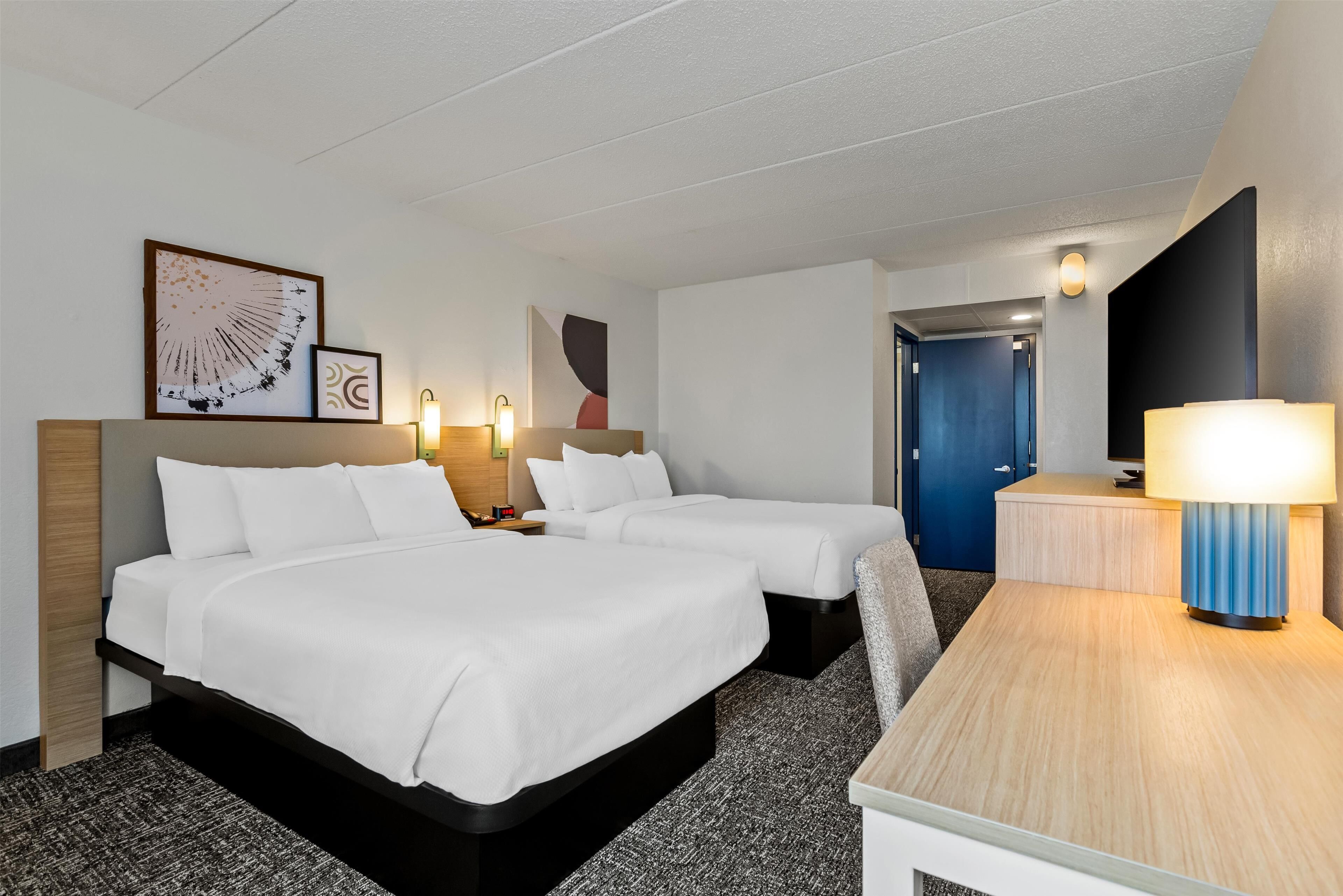 A hotel room with two beds, a desk, a lamp, and artwork on the wall.