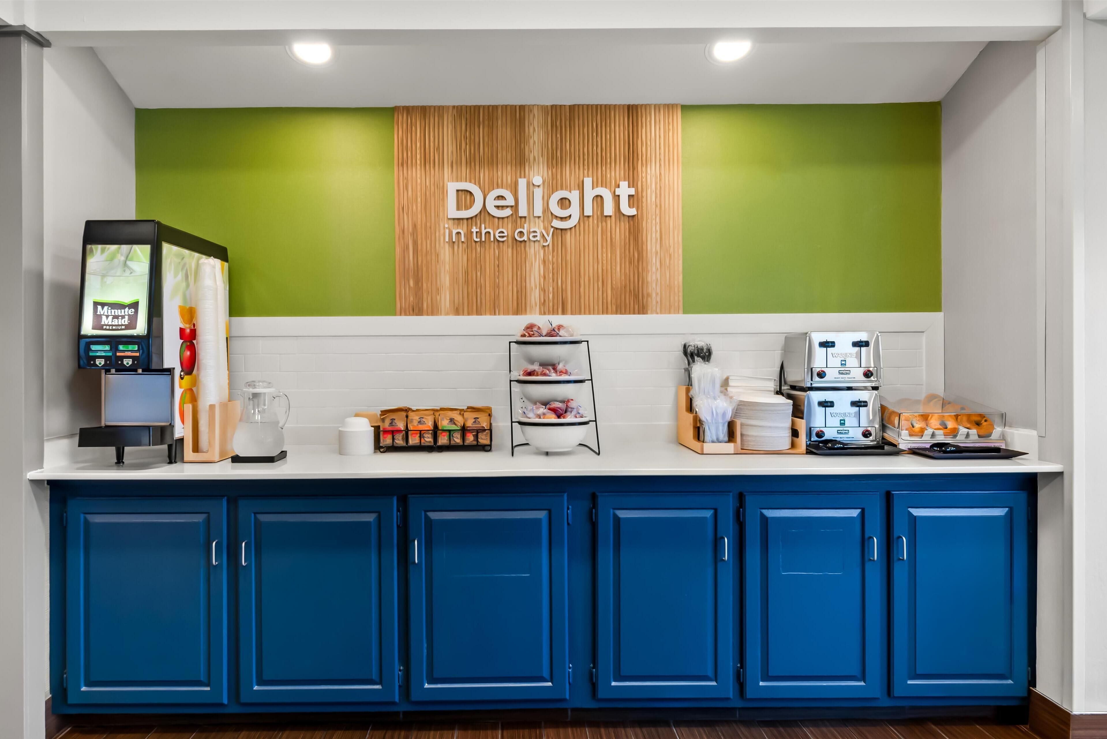 A hotel reception area with a green wall, a 'Delight in the day' sign, a self-service kiosk, and a buffet counter with various food items and a coffee machine.