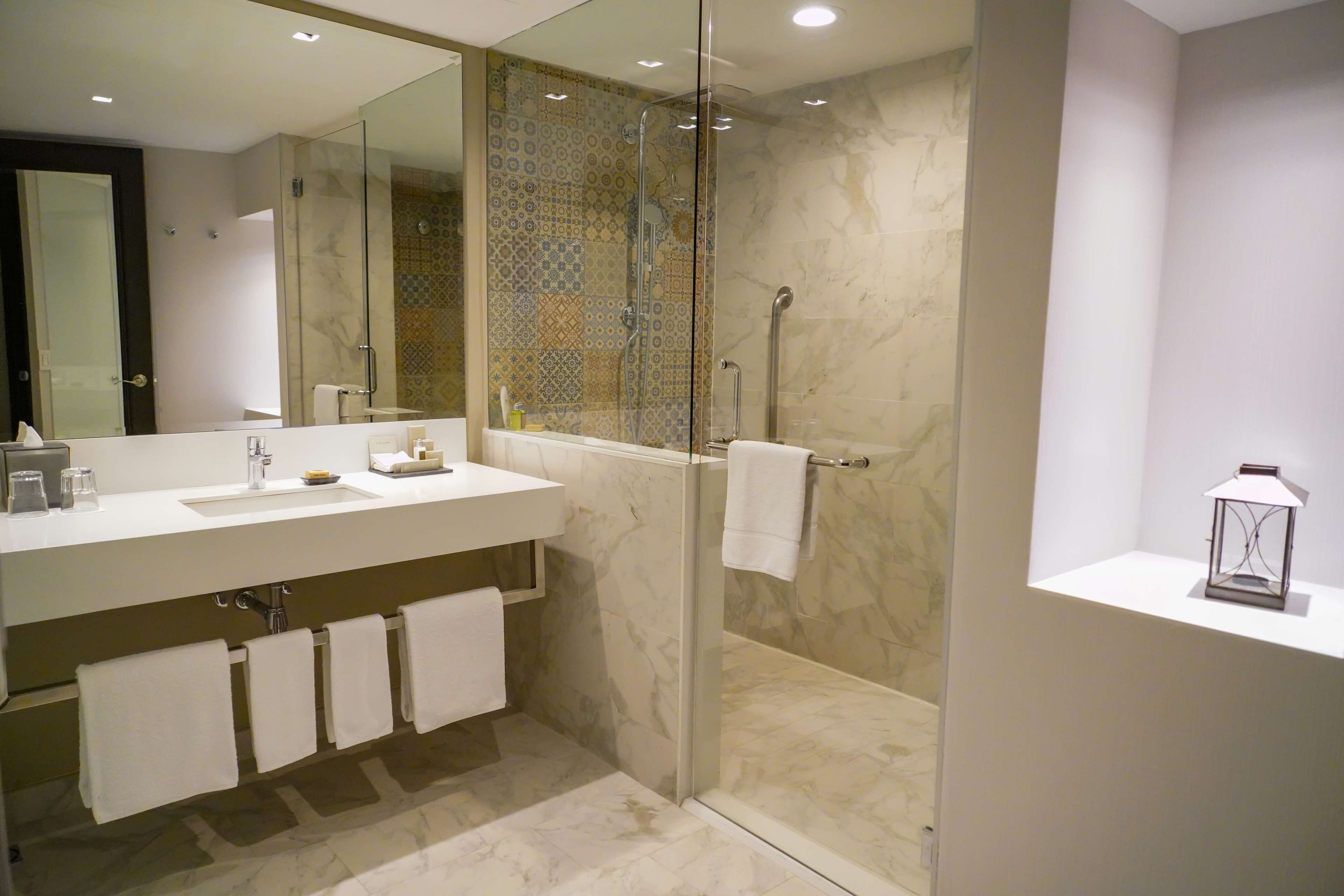 Un baño moderno con ducha envuelta en vidrio, un lavamanos blanco con espejo y un pequeño estante con un farol.
