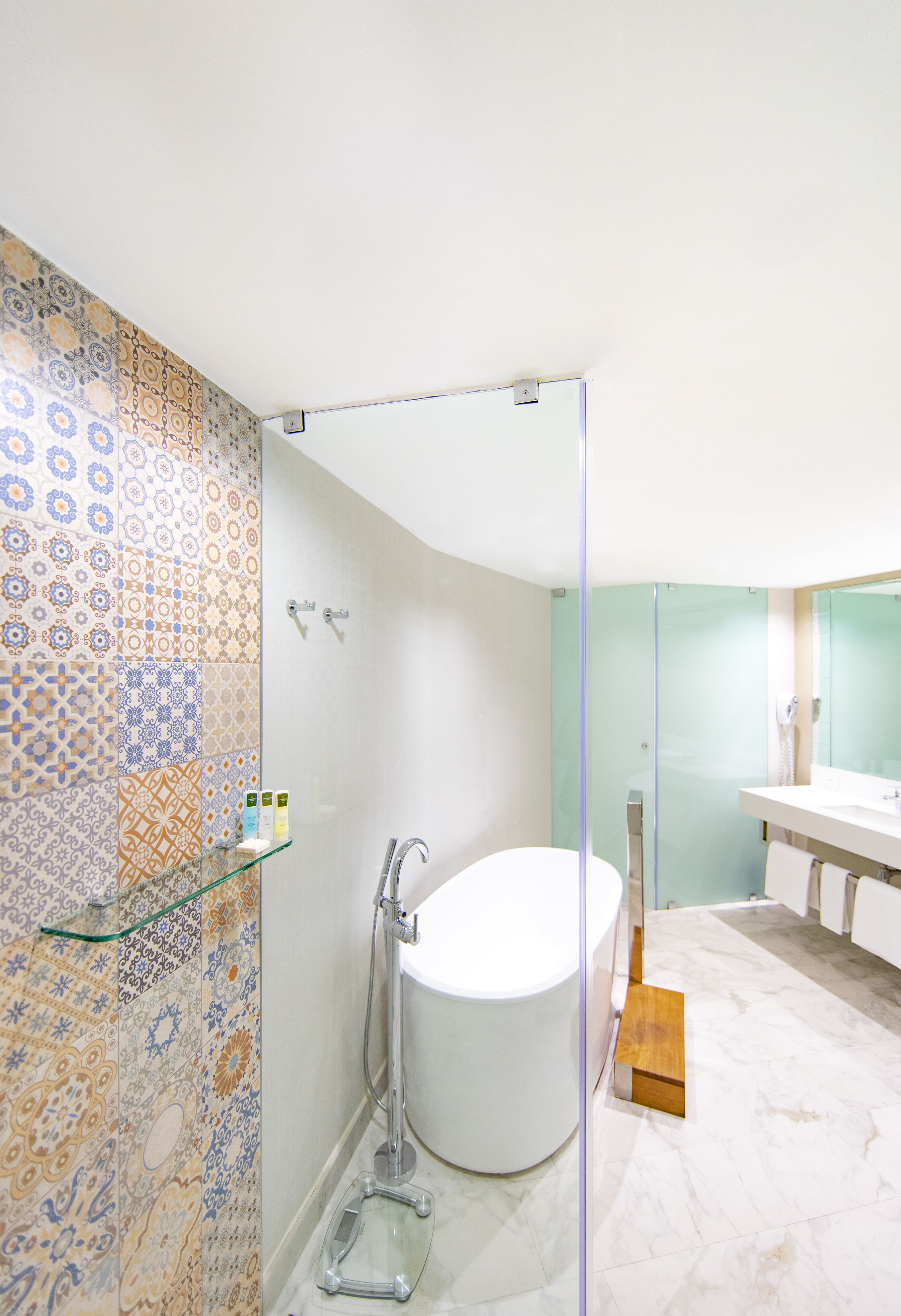 A modern bathroom with patterned tiles, a glass shower enclosure, a white bathtub, and a wooden step leading to the shower.