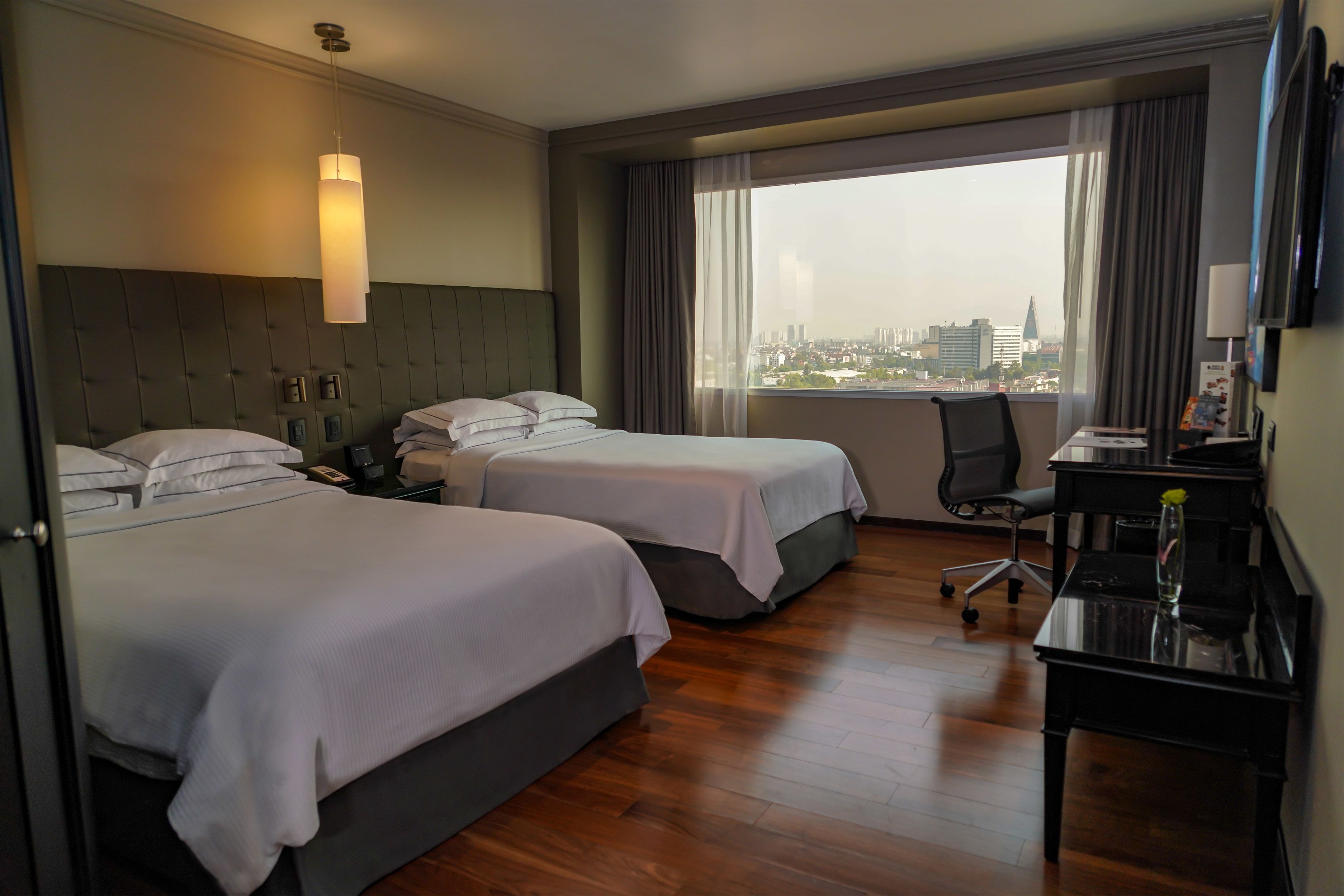 Une chambre d’hôtel avec deux lits, un bureau, une chaise et une grande fenêtre donnant sur la ville.