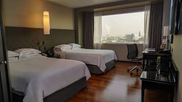 A hotel room with two beds, a desk, a chair, and a large window with a city view.