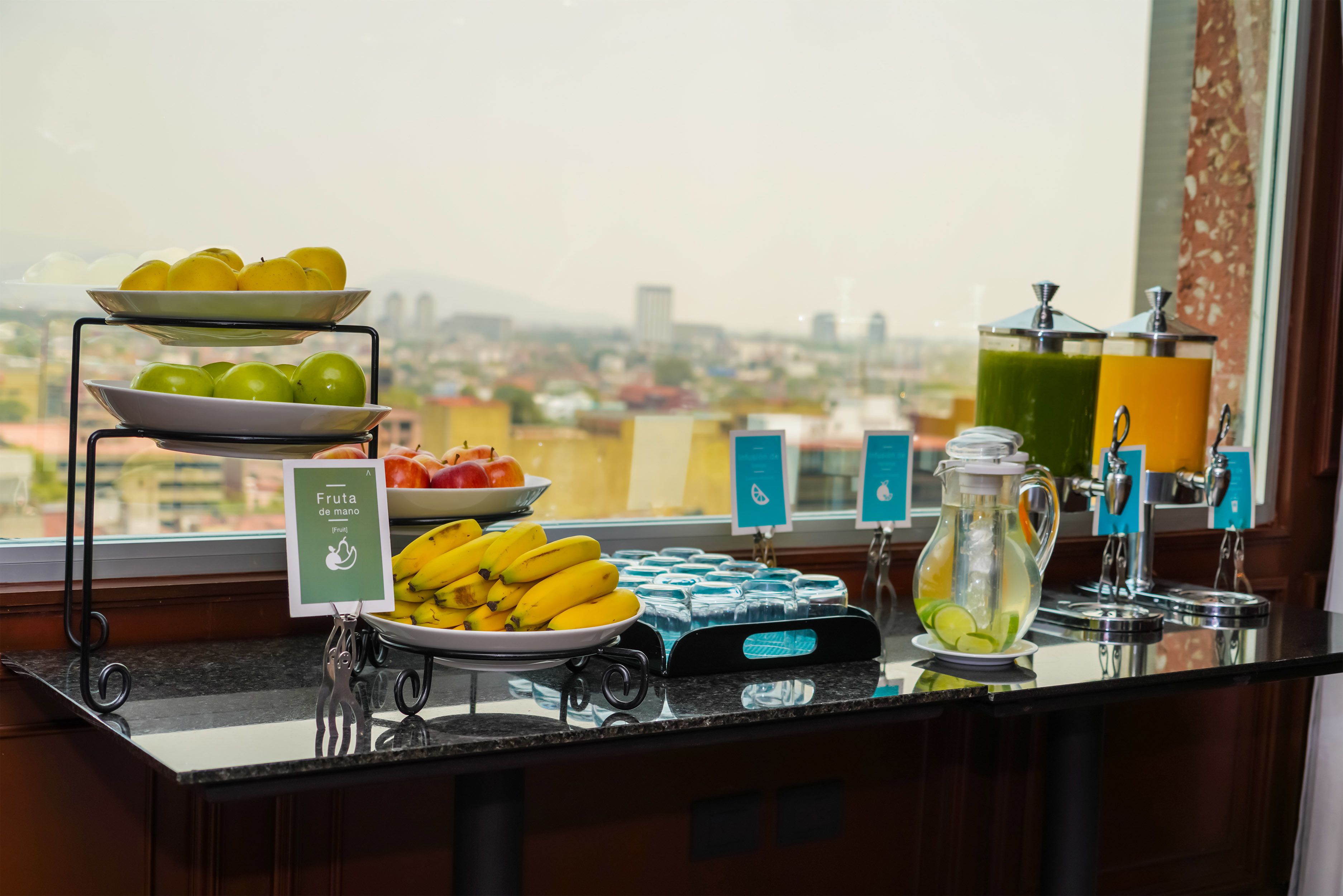 A buffet counter with various fruits and beverages, a city view in the background, and a sign indicating self-service.