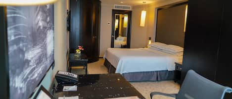Habitación de hotel con cama, mesita de noche, escritorio con papeles, una silla, lámpara y un espejo grande.