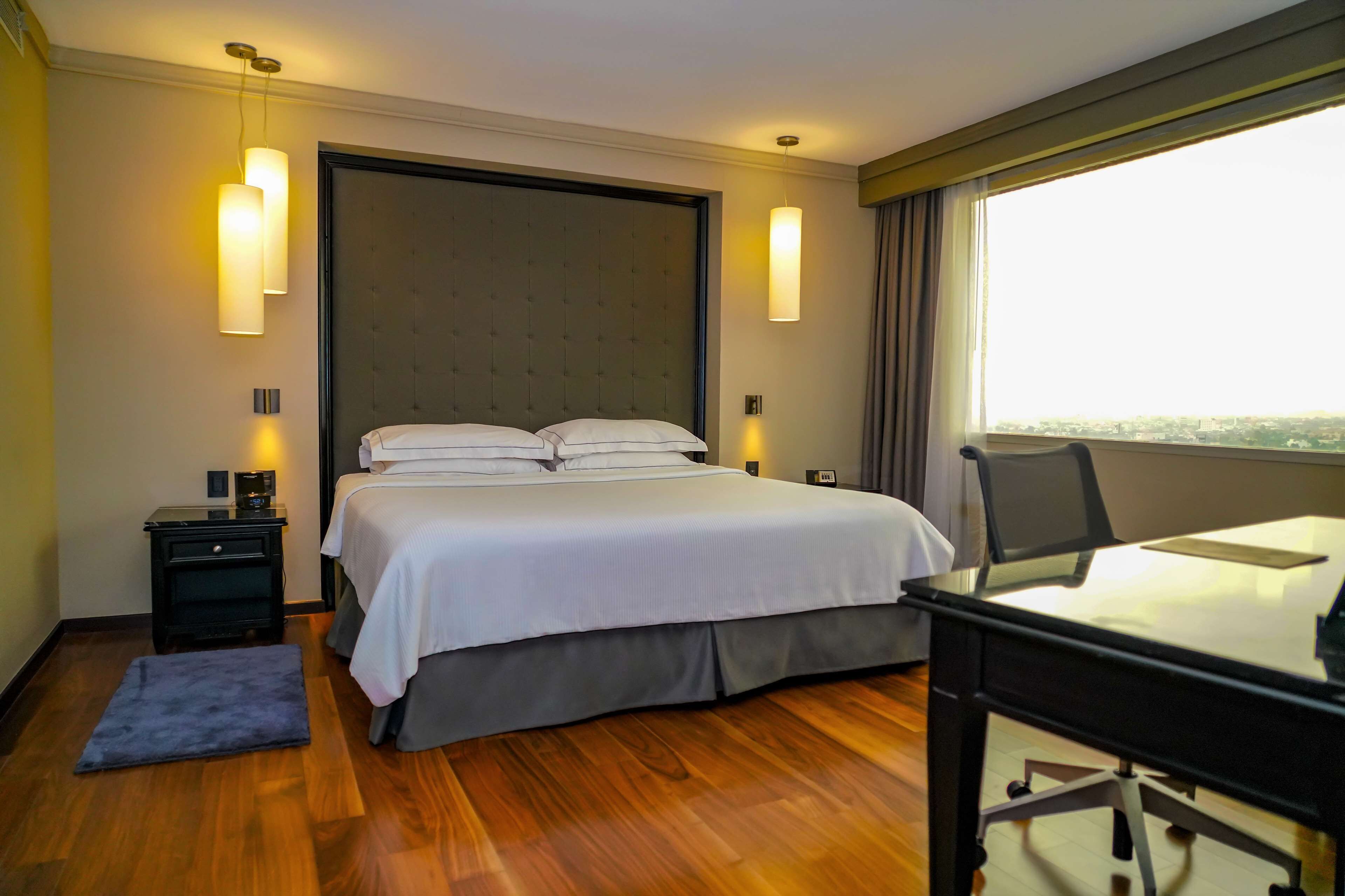Habitación de hotel con cama, mesitas de noche, un escritorio y vistas a la ciudad.