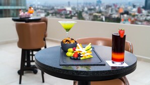 A table with a fruit platter, a martini glass, and a cocktail with a strawberry garnish, set against a cityscape view.