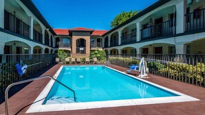 Una piscina con un paseo perimetral, un edificio con varios balcones y un cielo azul despejado.