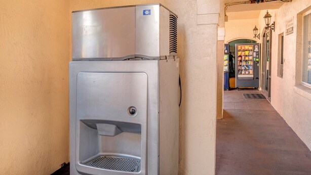 Un dispensador metálico compacto para bebidas con función de enfriador de agua, situado en un pasillo con pared texturizada y una máquina expendedora al fondo.