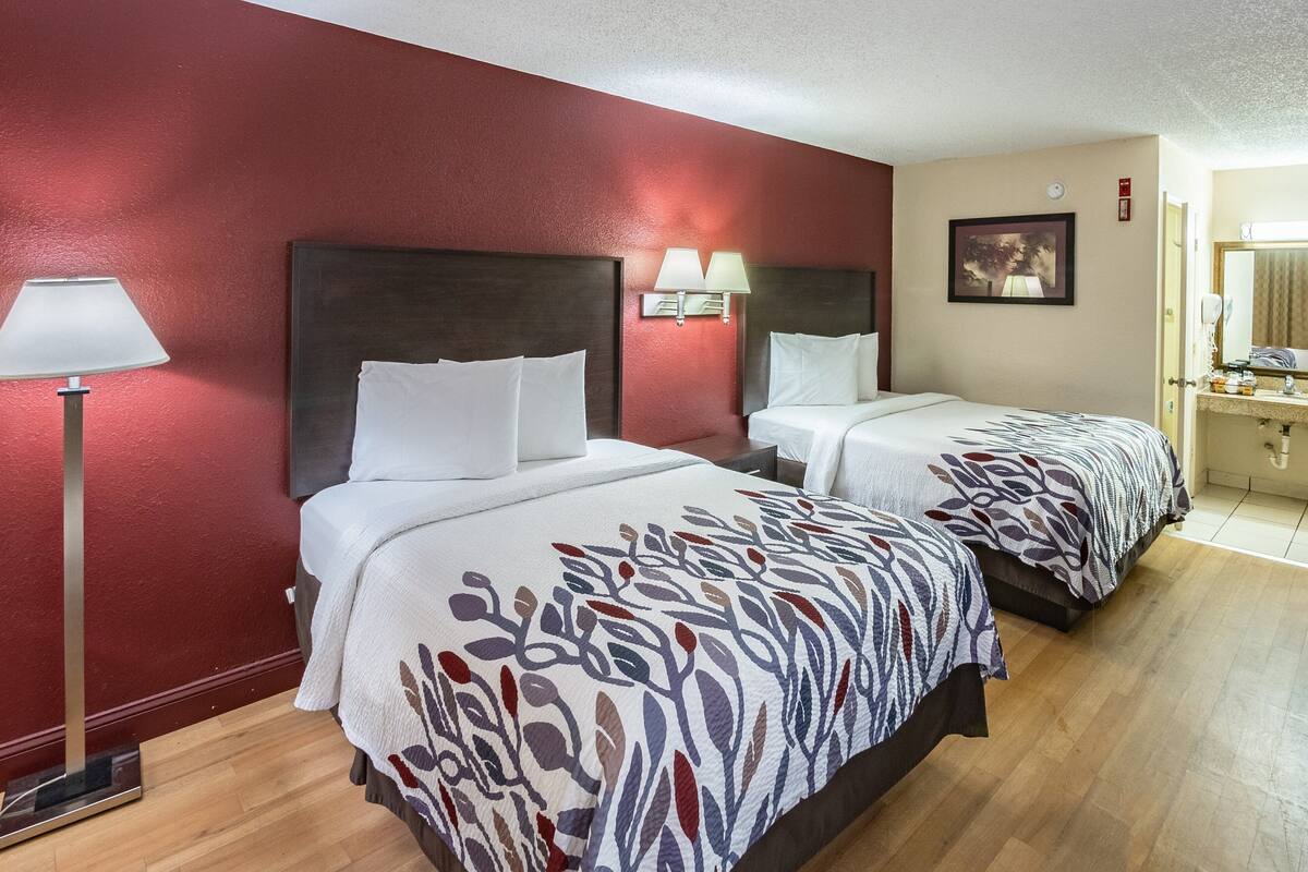 Una habitación de hotel con dos camas, una mesita de noche, una lámpara y un baño con lavamanos y espejo.