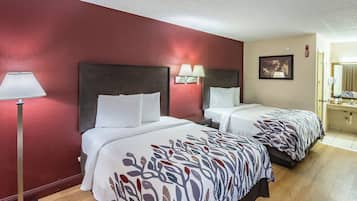 A hotel room with two beds, a nightstand, a lamp, a painting, and a bathroom with a sink and mirror.
