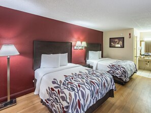 Habitación de hotel con dos camas, una mesita de noche, una lámpara, un cuadro y un baño con lavamanos y espejo.