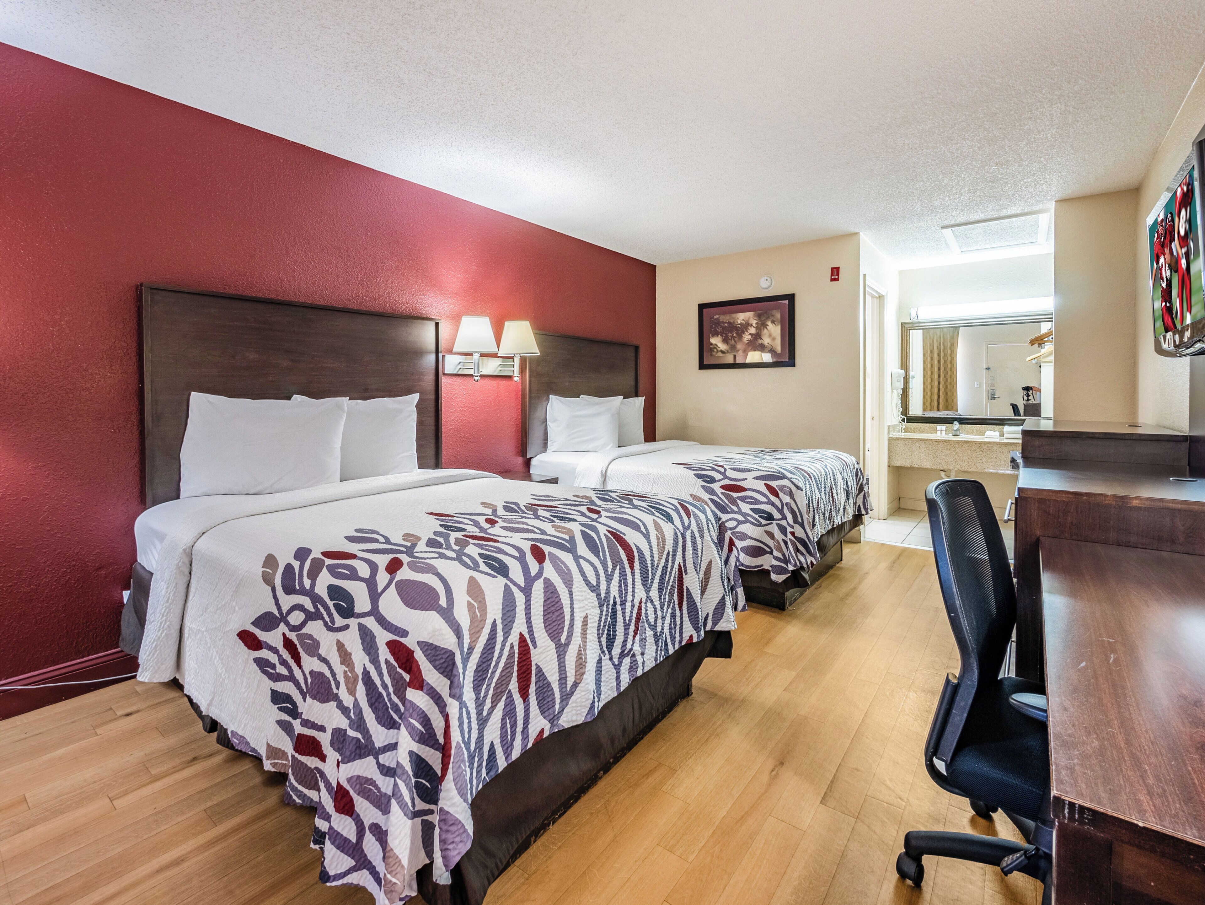 A hotel room with two beds, a desk, a chair, a mirror, and a window.