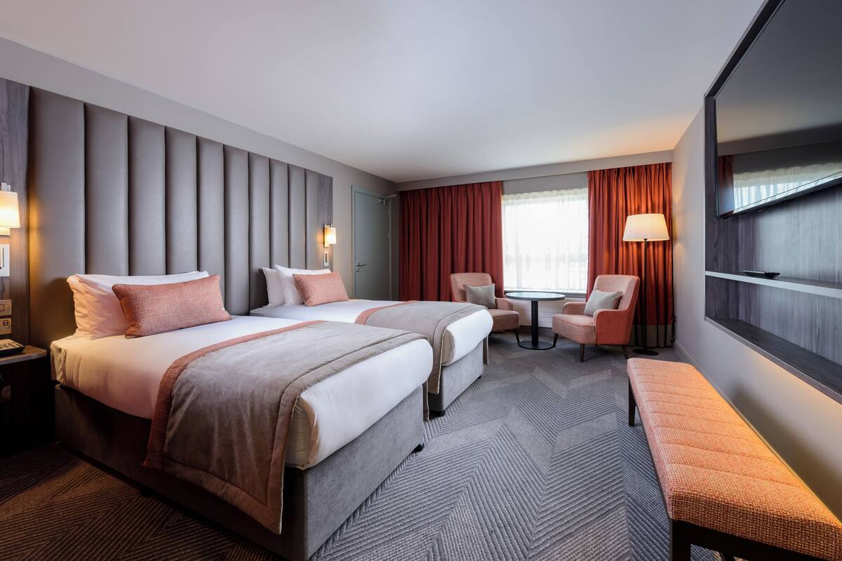 A hotel room with two beds, a desk, a chair, a bench, and a TV.