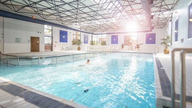 An indoor swimming pool with clear water, a metal roof structure, and several chairs along the poolside. - Hotel Killarney (Killarney)