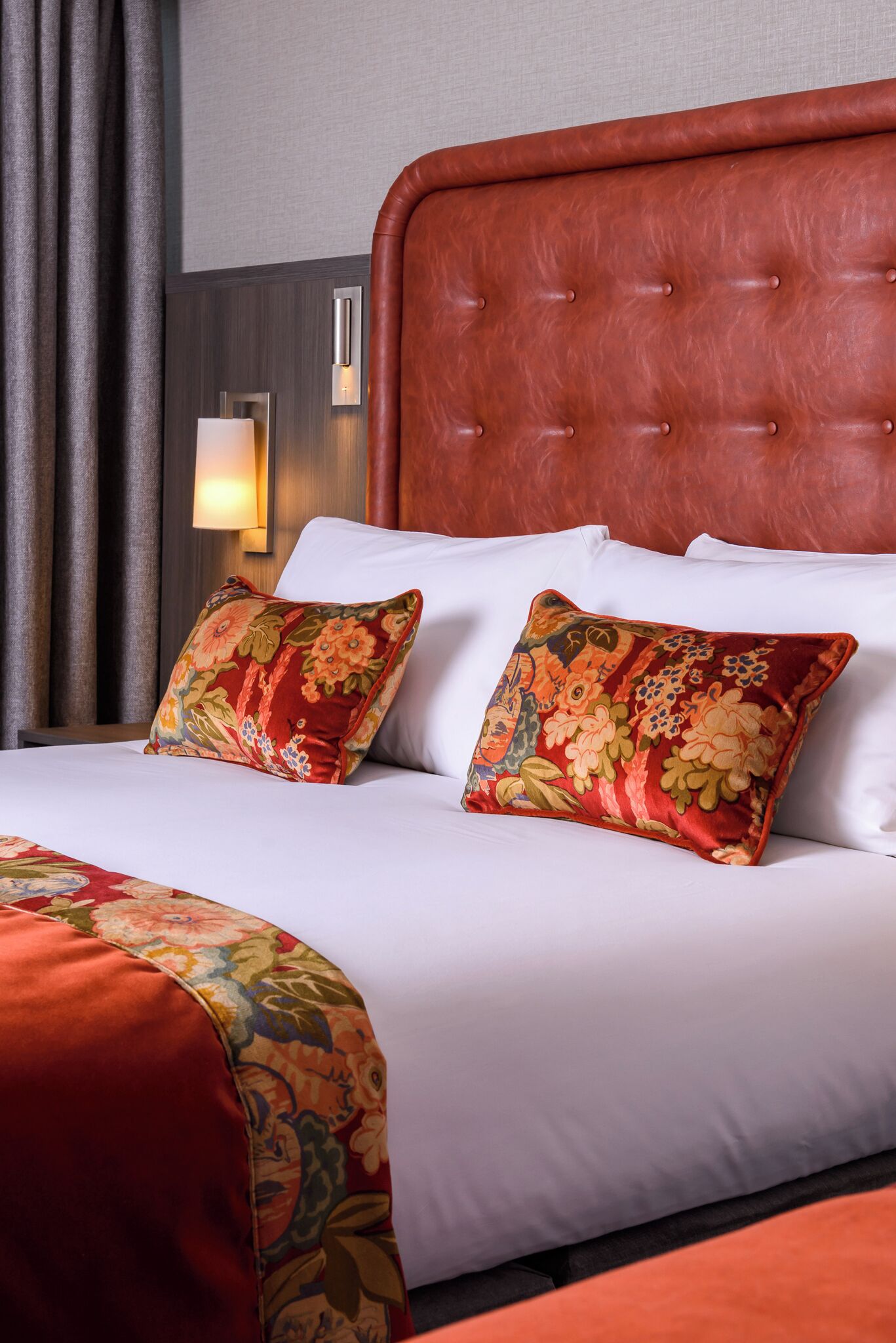 A hotel room with a large bed, two pillows with floral patterns, a red headboard, and a wall-mounted light.