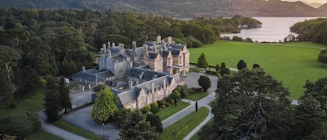 A large, historic building with multiple chimneys, surrounded by lush greenery and a lake.