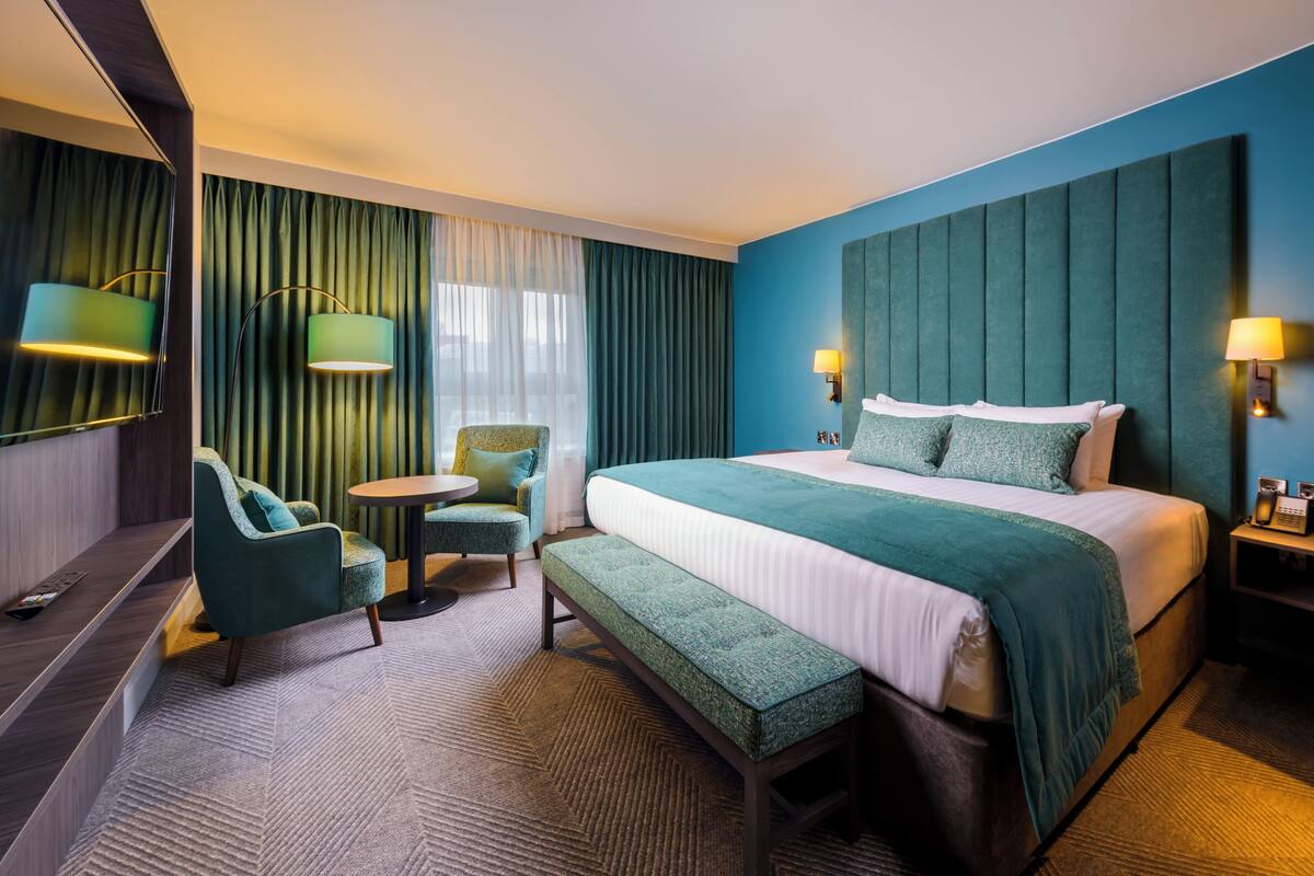 A hotel room with a large bed, a bench, a chair, a small table, and a mirror.