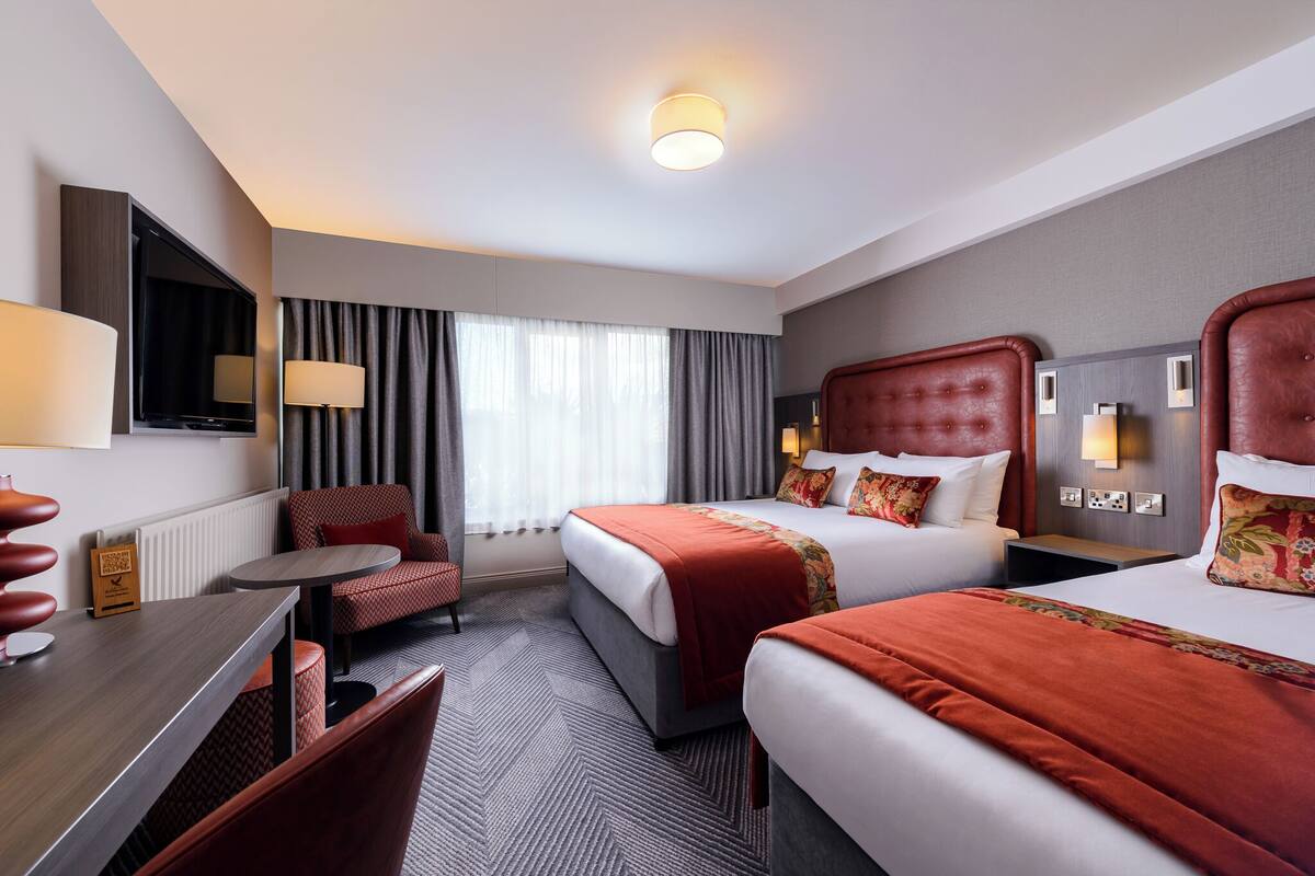 A hotel room with two beds, a desk, a chair, a TV, and a window with curtains.