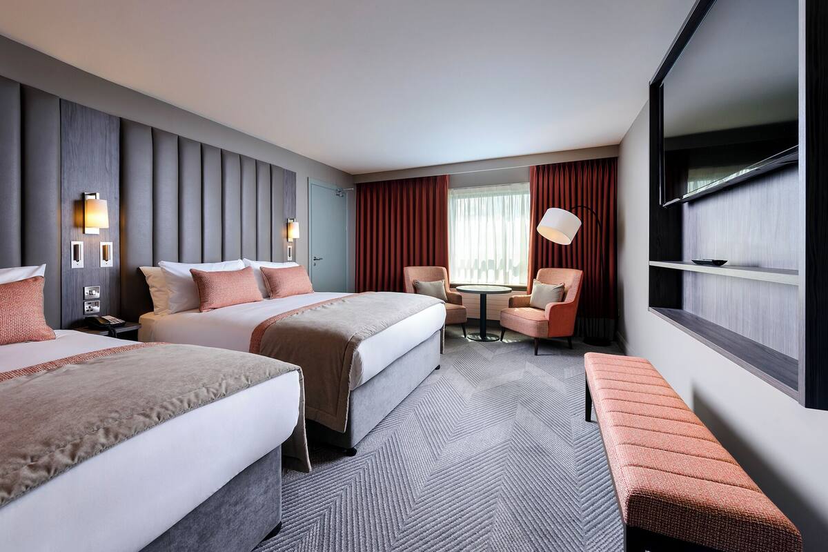 A hotel room with two beds, a small round table, a bench, and a large window with curtains.