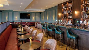 Una zona de bar con diversas opciones de asientos, incluyendo taburetes y mesas redondas, una barra con una variedad de botellas de licores y una pantalla de televisión en la pared.