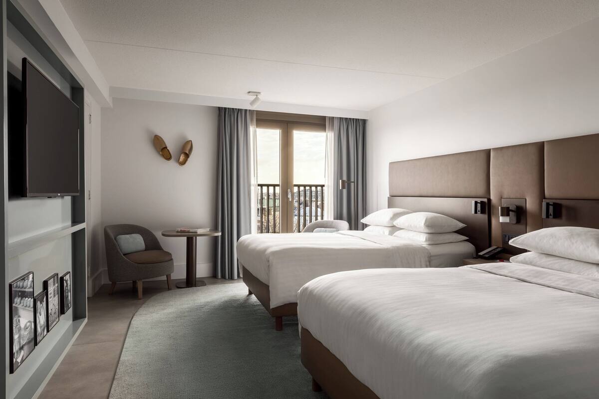 Une chambre d’hôtel avec deux lits, une télévision, une petite table, une chaise et une vue sur la ville par la fenêtre.