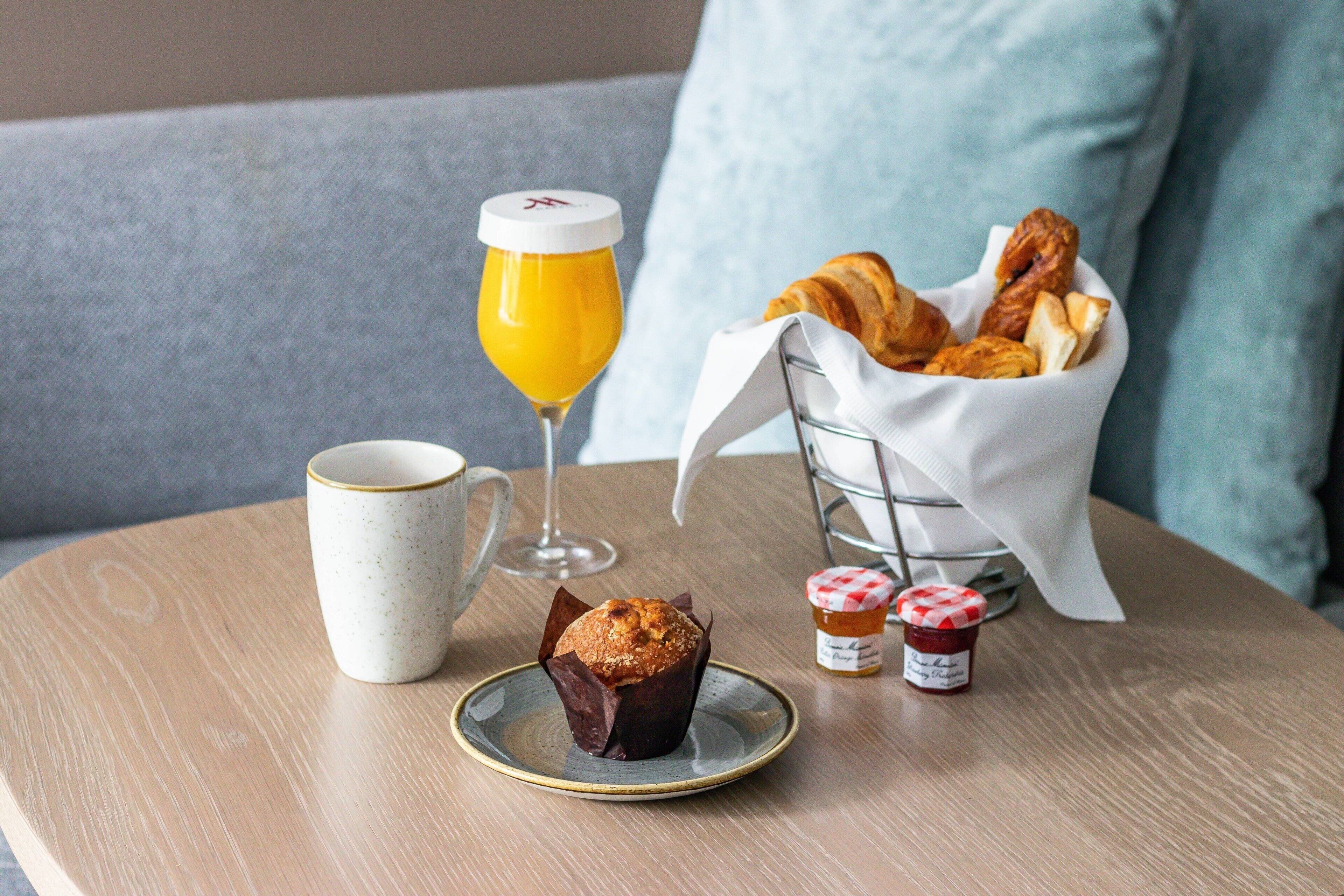 A breakfast setting with a muffin, croissants, a glass of orange juice, and two jars of jam on a wooden table.
