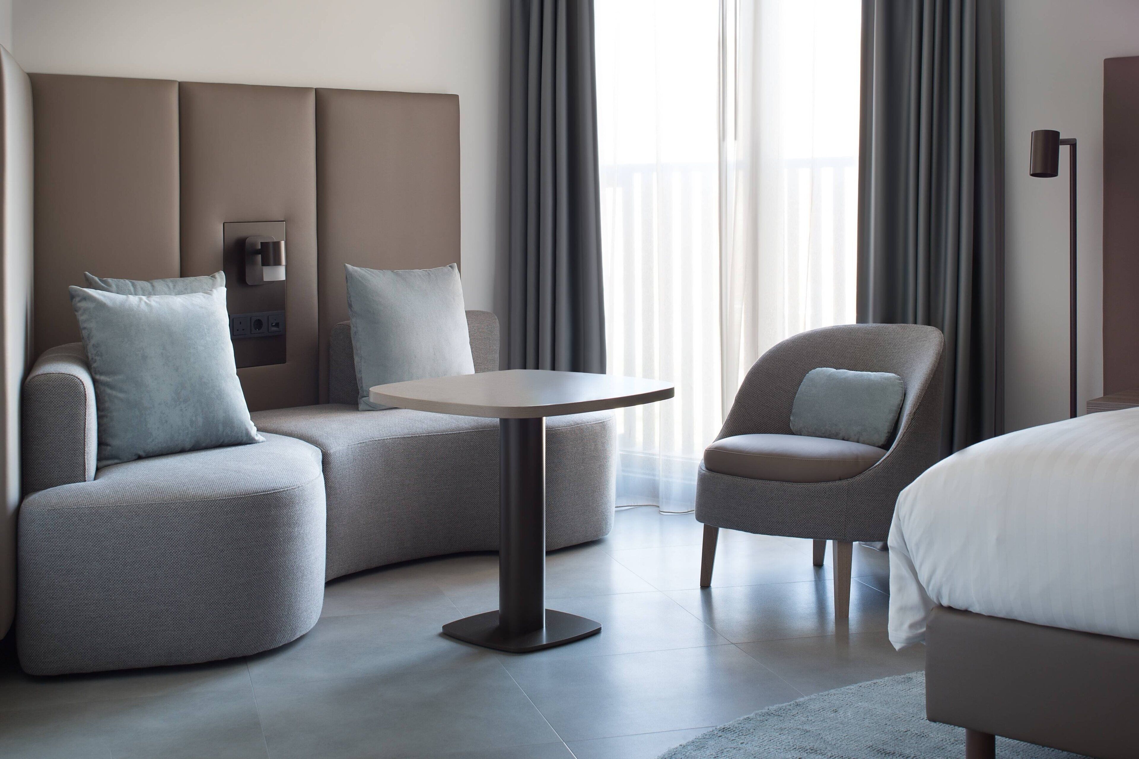 A modern hotel room with a curved sofa, a round table, a single armchair, and a bed.