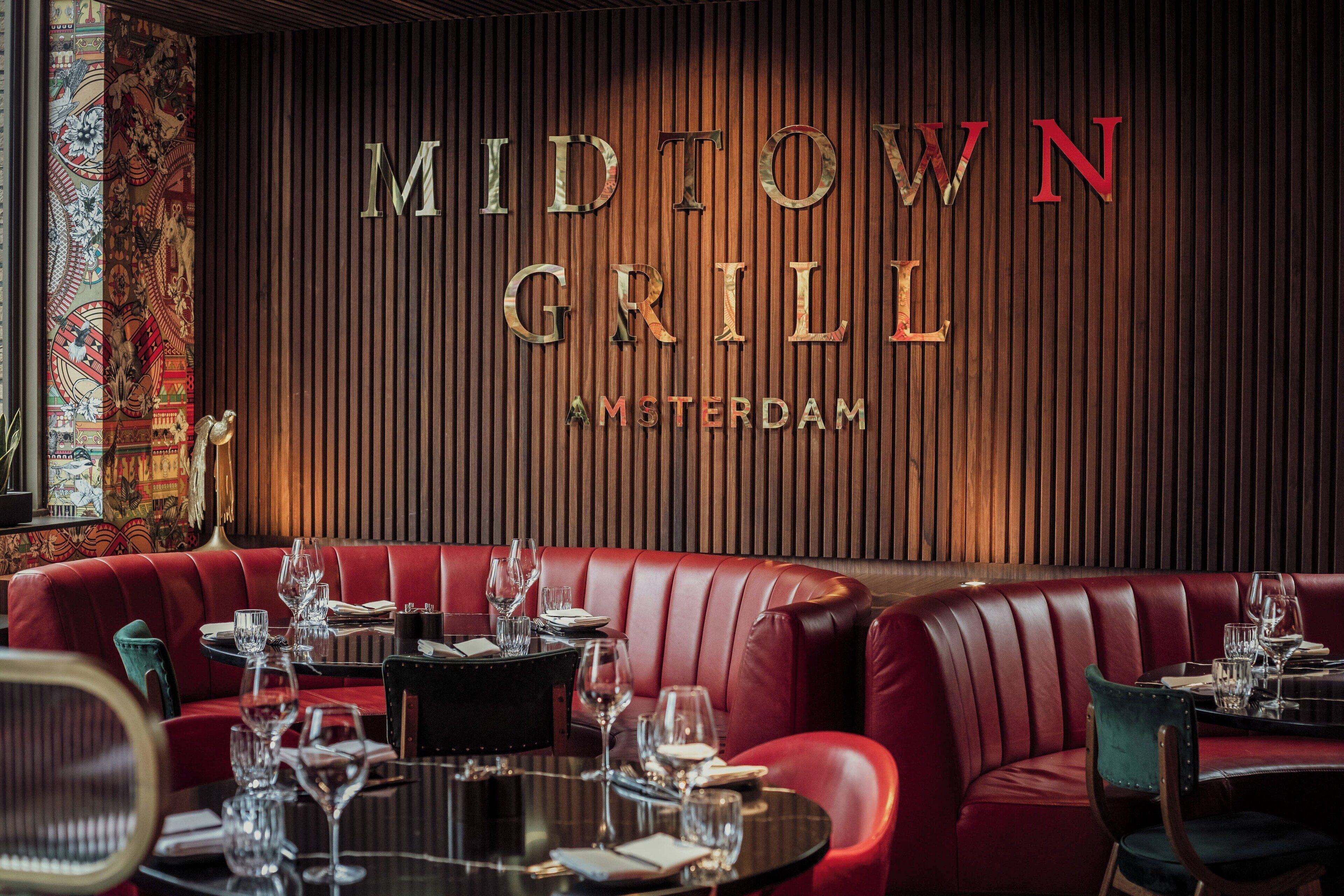 A restaurant interior with red leather seating, a wooden wall with the name 'MIDTOWN GRILL AMSTERDAM' in large letters, and a patterned tapestry on the left.