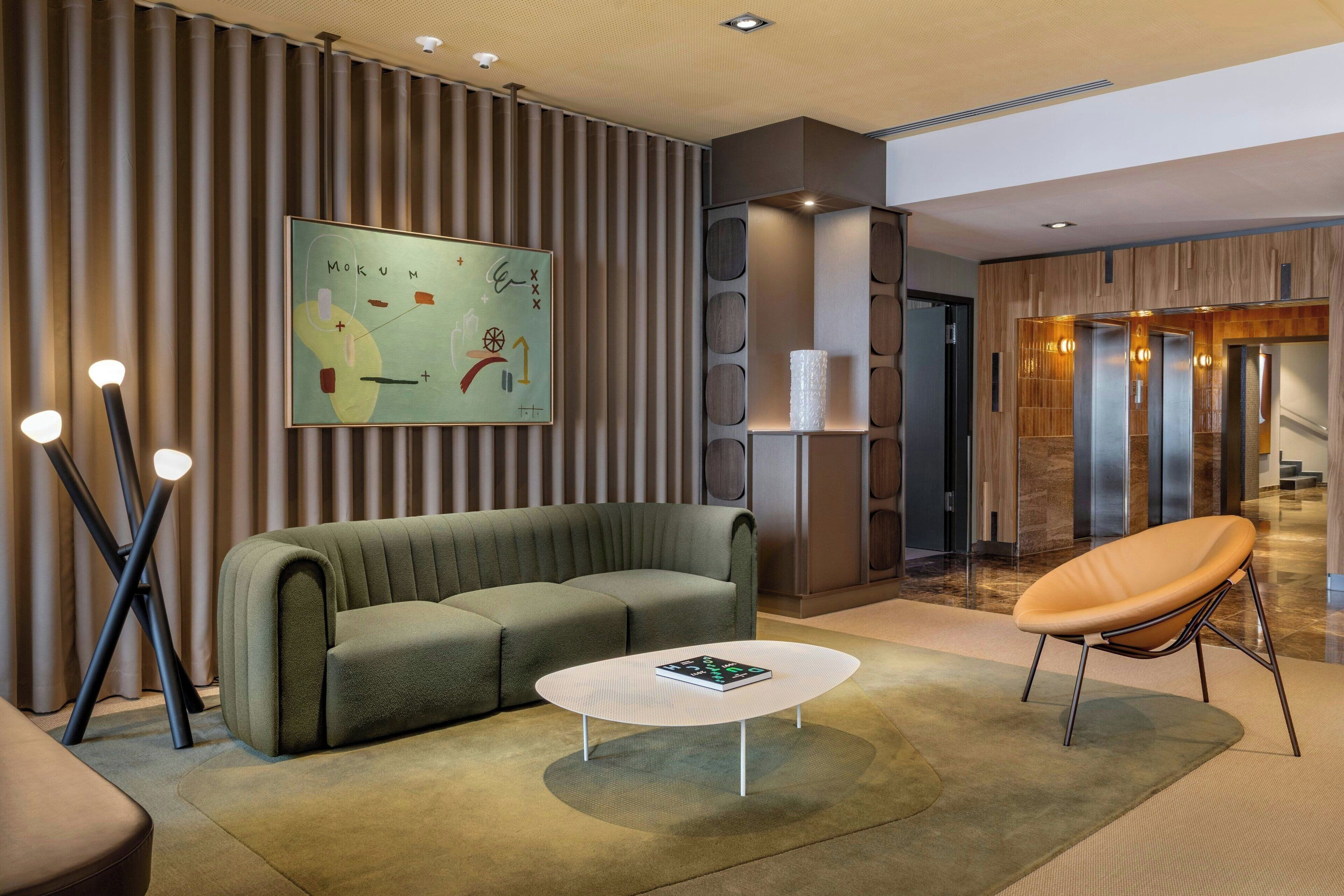 A modern lobby with a sectional sofa, a coffee table, and a lounge chair. There is a wall-mounted artwork and a reception desk with a glass partition.