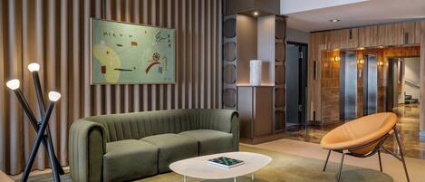 A modern lobby with a sectional sofa, a coffee table, and a lounge chair. There is a wall-mounted artwork and a reception desk with a glass partition.
