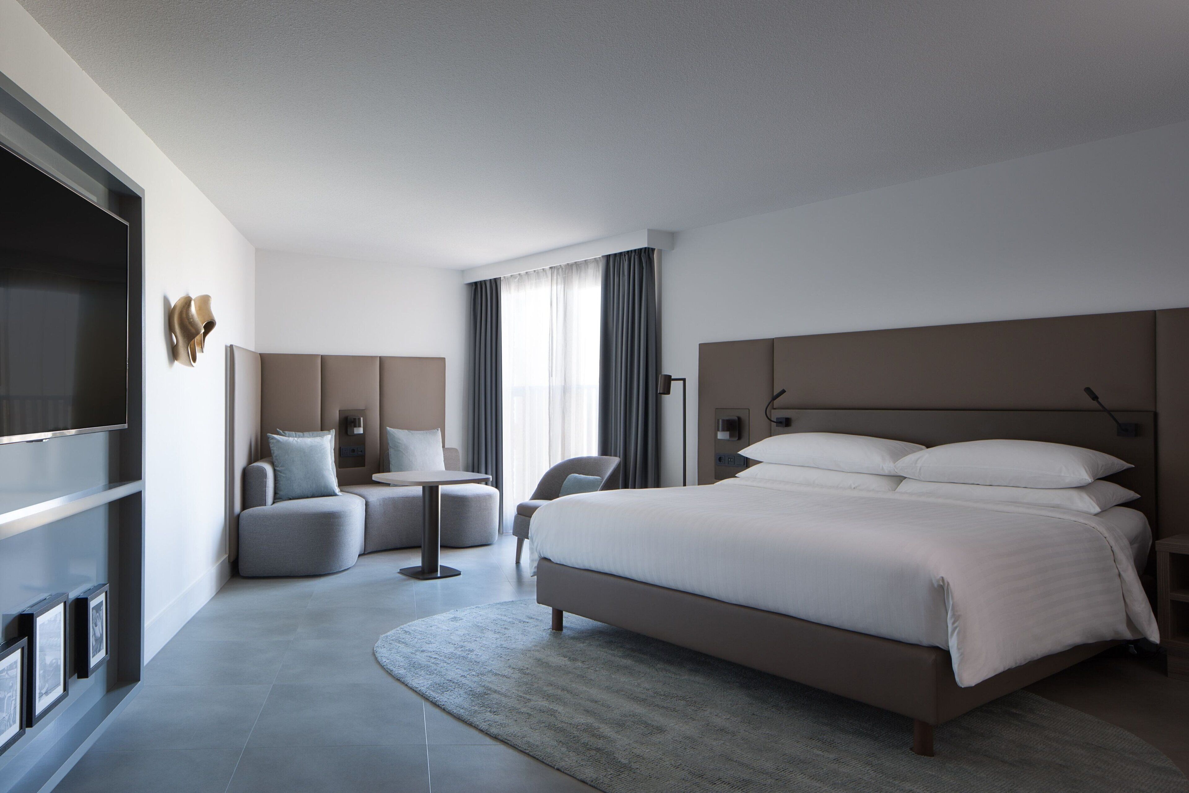 A modern hotel room with a large bed, a flat-screen TV, a seating area with a table, and a large window with curtains.