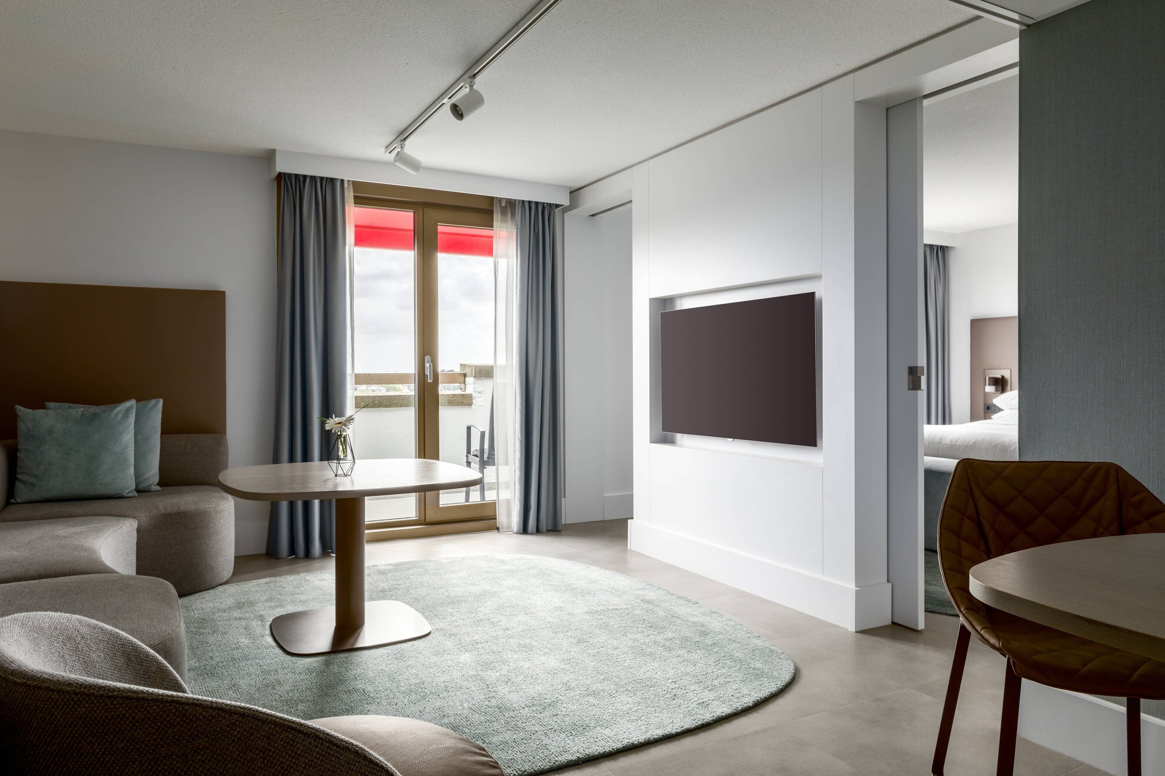 A modern hotel room with a sofa, a round table, a TV, and a balcony with curtains.