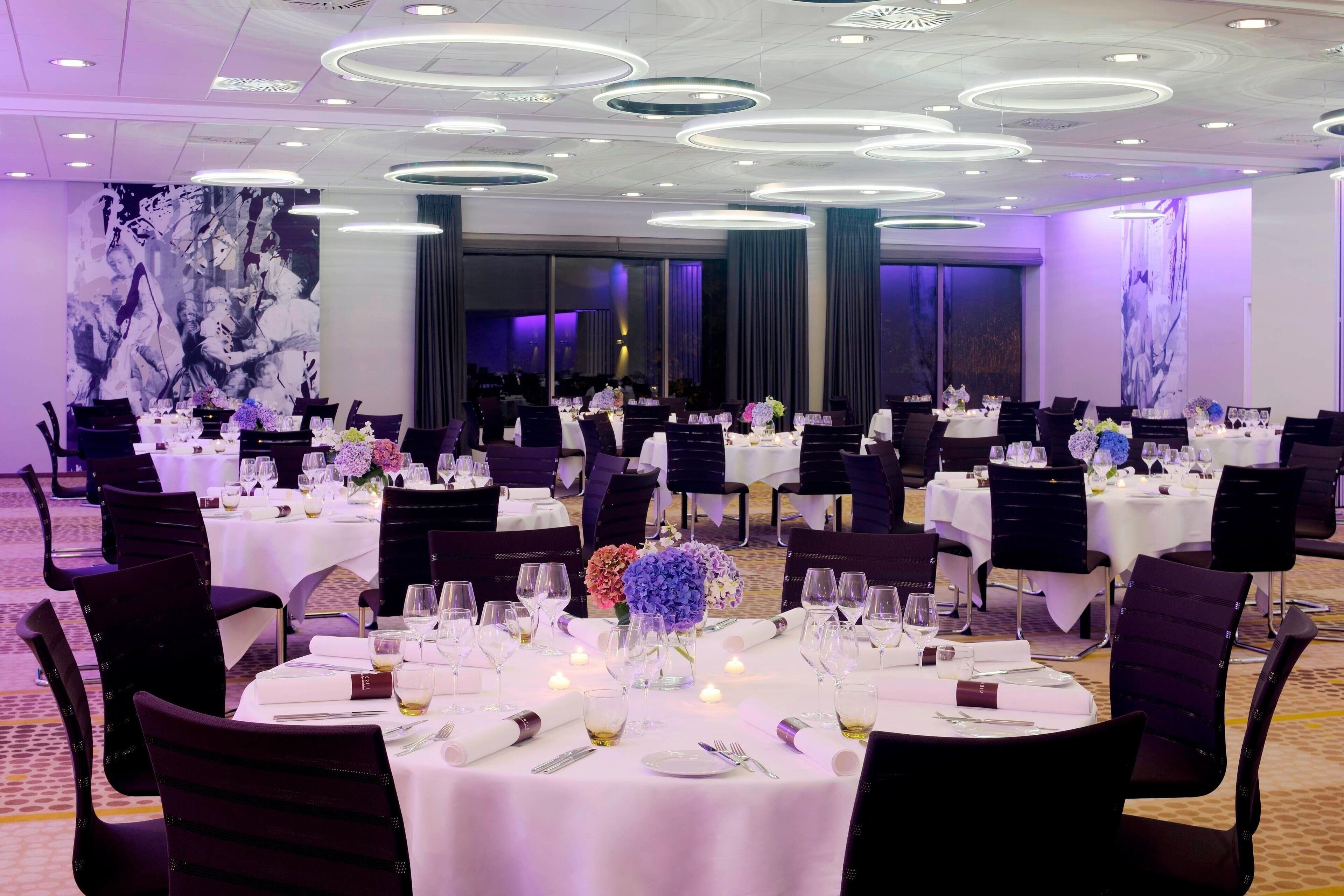 A banquet hall with round tables set for dining, featuring white tablecloths, dark chairs, and floral centerpieces.