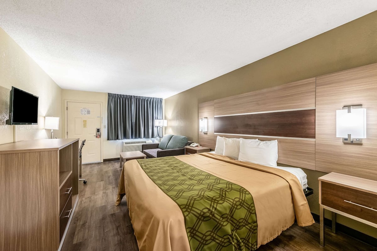 A hotel room with a bed, bedside table, lamp, TV, and a desk with a chair.