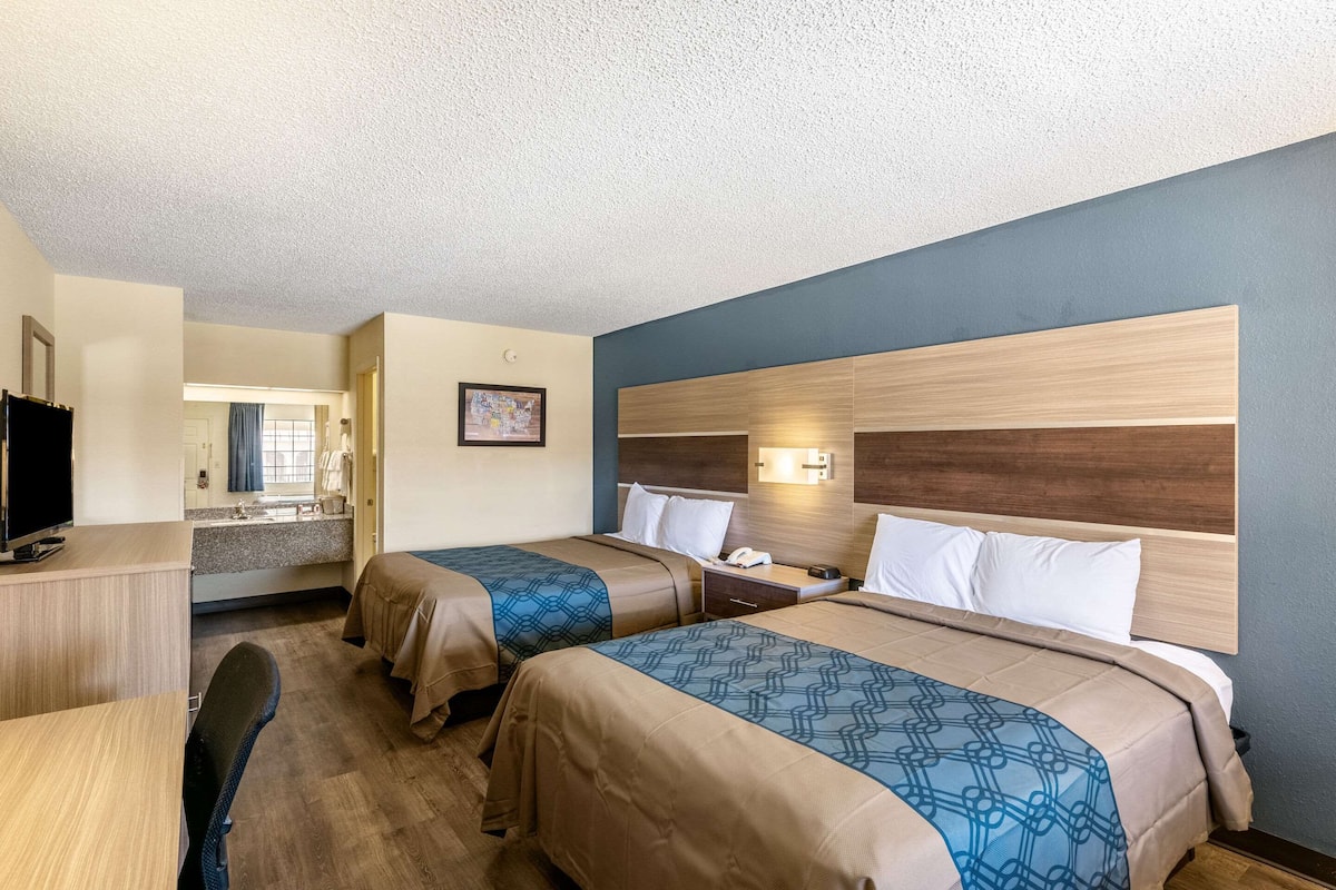 A hotel room with two beds, a desk, a chair, a small kitchenette, and a bathroom visible through an open door.