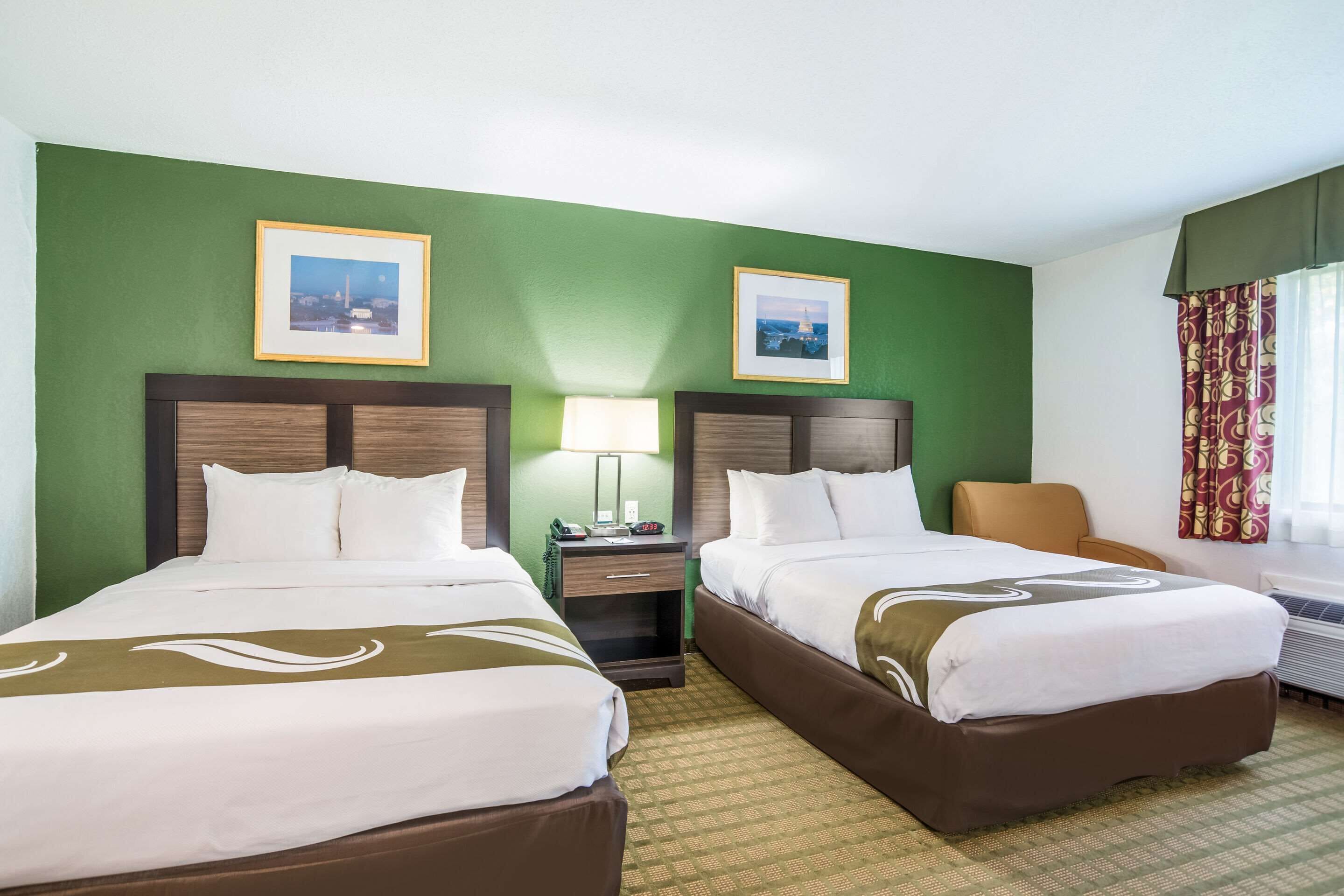 A hotel room with two beds, green walls, framed pictures, a nightstand, a lamp, and a window with curtains.