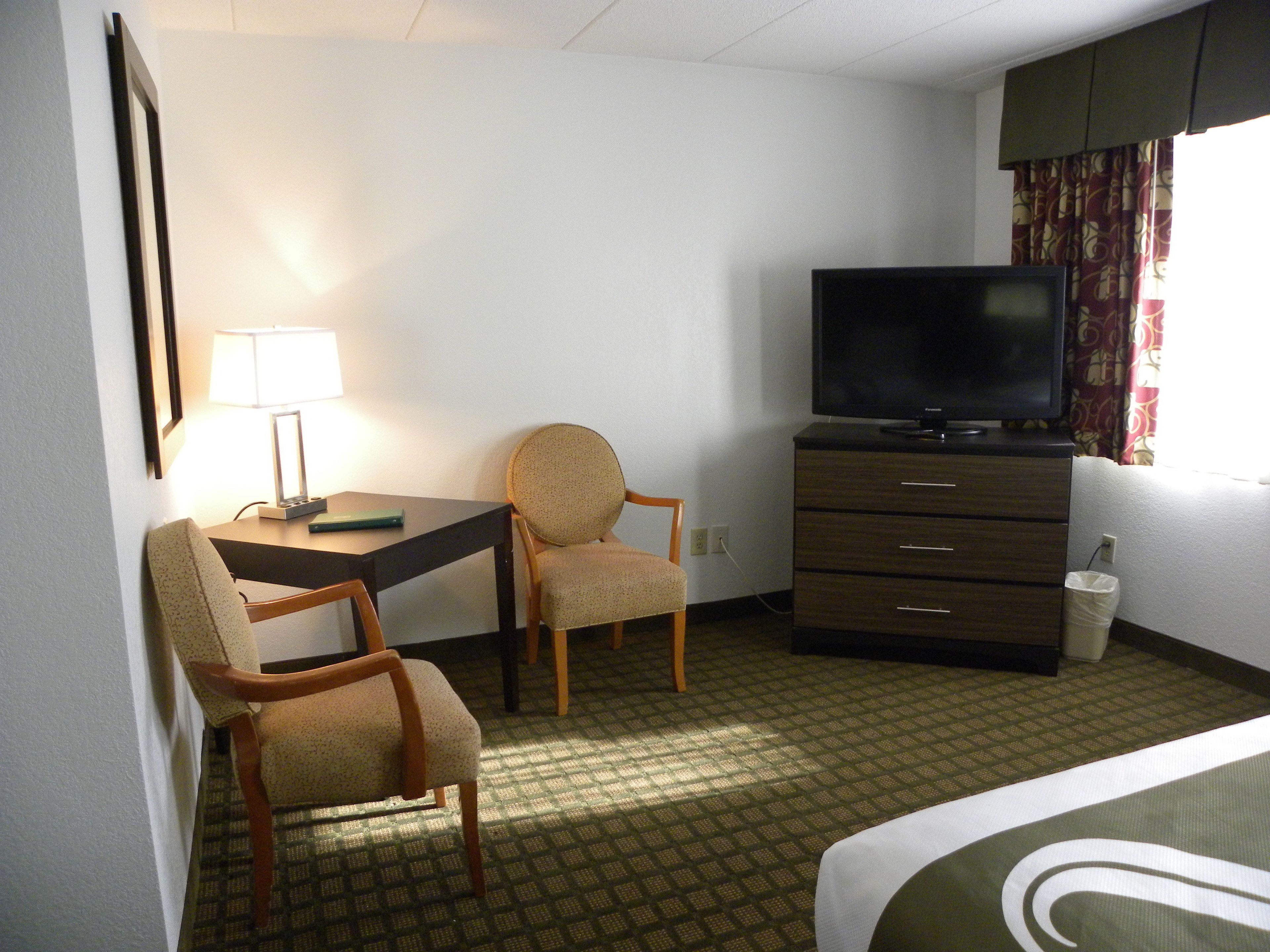 A hotel room with a bed, a desk with a chair, a television, a lamp, and a waste bin.