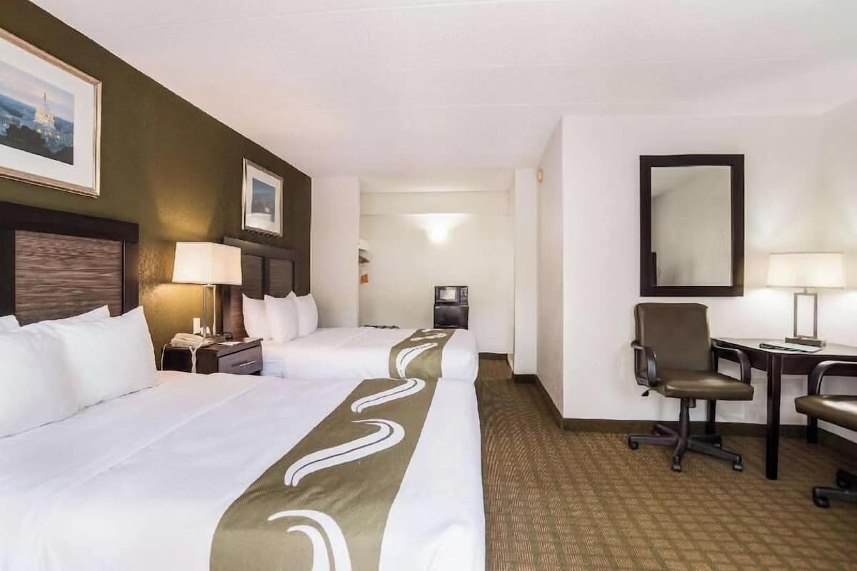 Habitación de hotel con dos camas, un escritorio, una silla, un espejo y una mesita de noche con lámpara.
