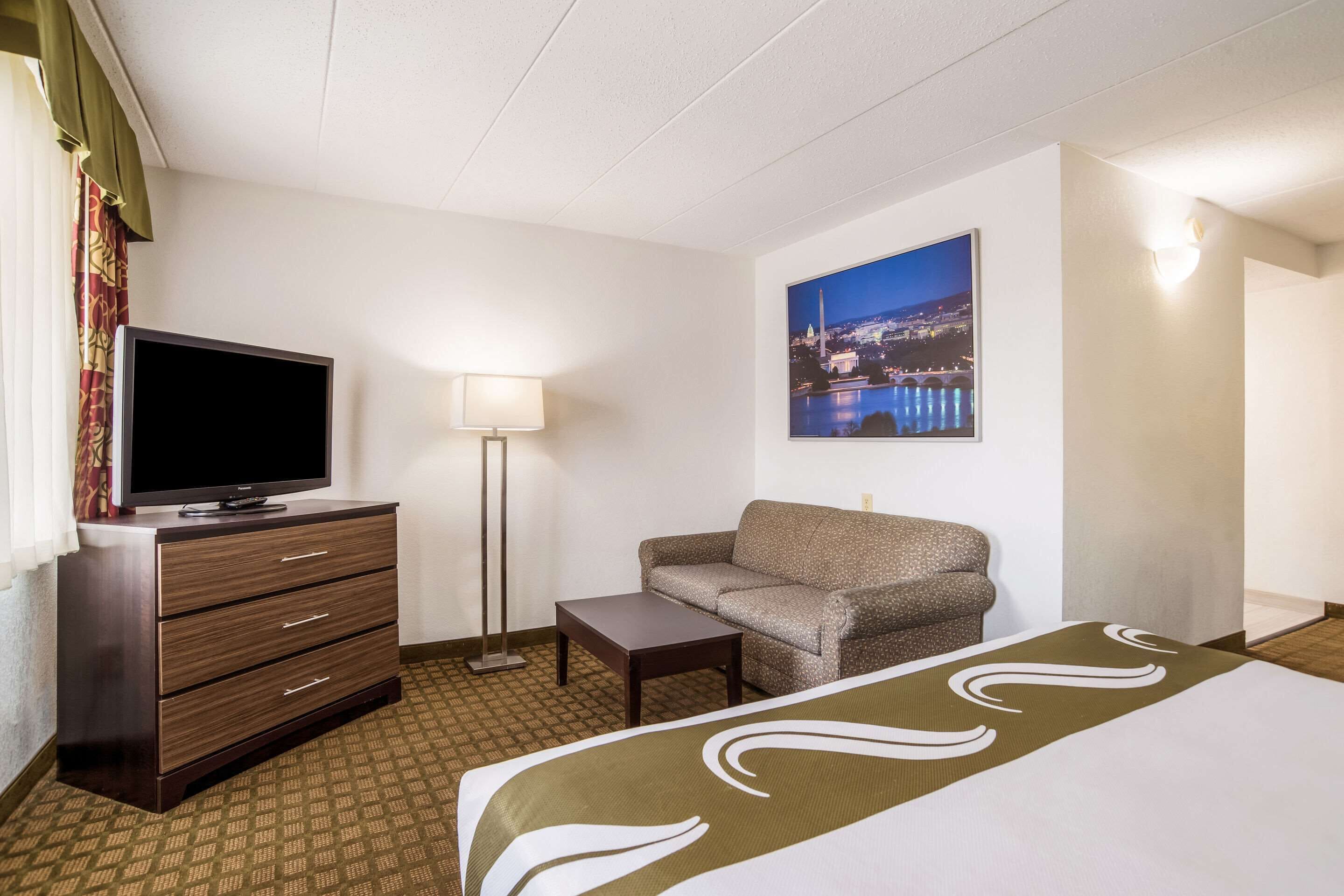 A hotel room with a bed, a television on a dresser, a sofa, a small table, and a painting of a cityscape.