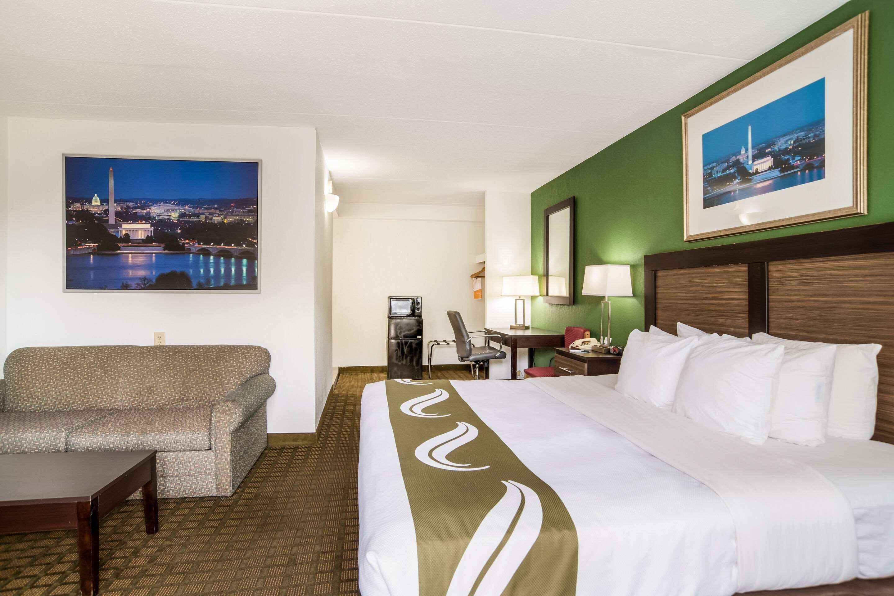 Ein Hotelzimmer mit einem großen Bett, einer Sitzecke, einem Schreibtisch mit Computer und zwei gerahmten Bildern an den Wänden.