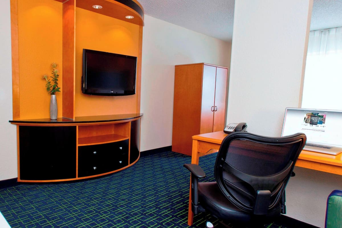 Una habitación de hotel con un escritorio curvo, una silla de oficina negra, un televisor montado en la pared y un armario.