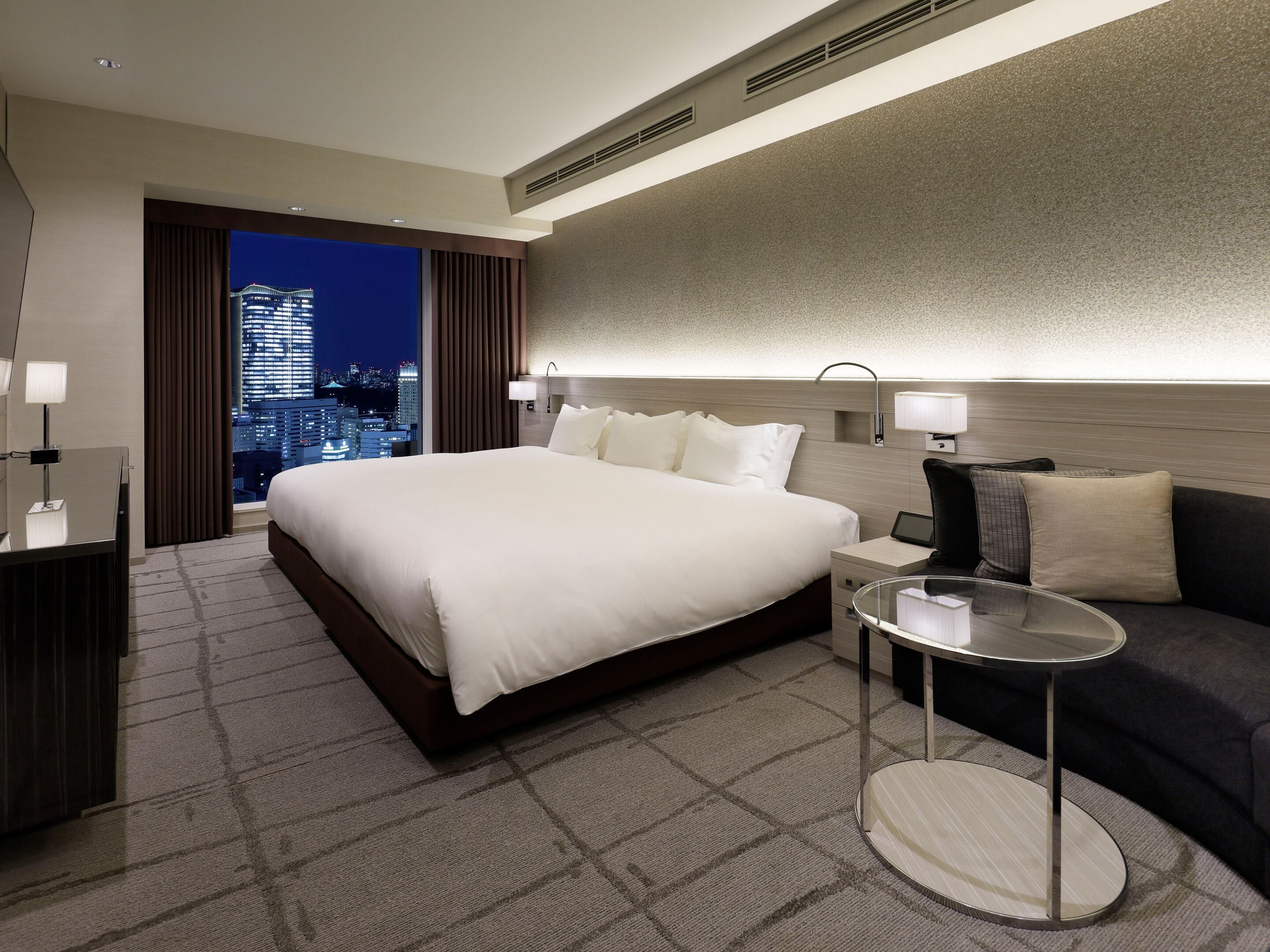 A hotel room with a large bed, a desk, a chair, a round table, and a view of the cityscape at night.