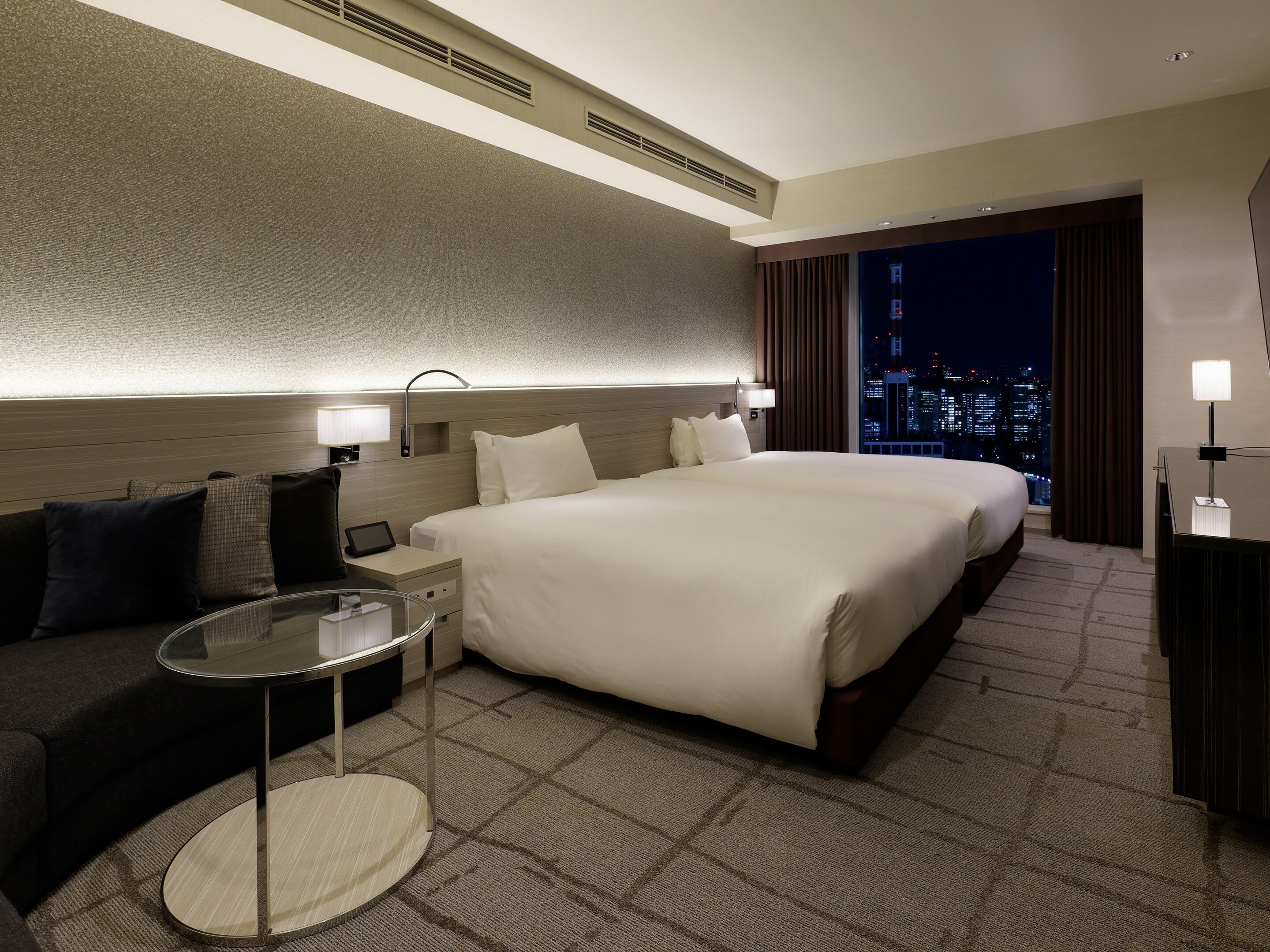 A hotel room with a large bed, a small desk, a round table, a sofa, and a view of the city at night.