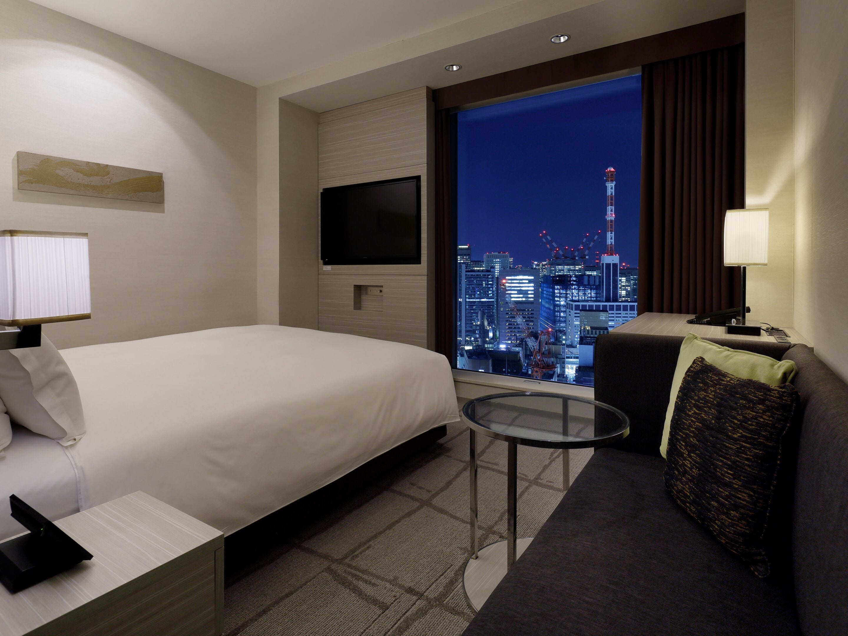 Une chambre d’hôtel avec un lit, une petite table, un canapé, une télévision et une vue sur le paysage urbain illuminé la nuit.
