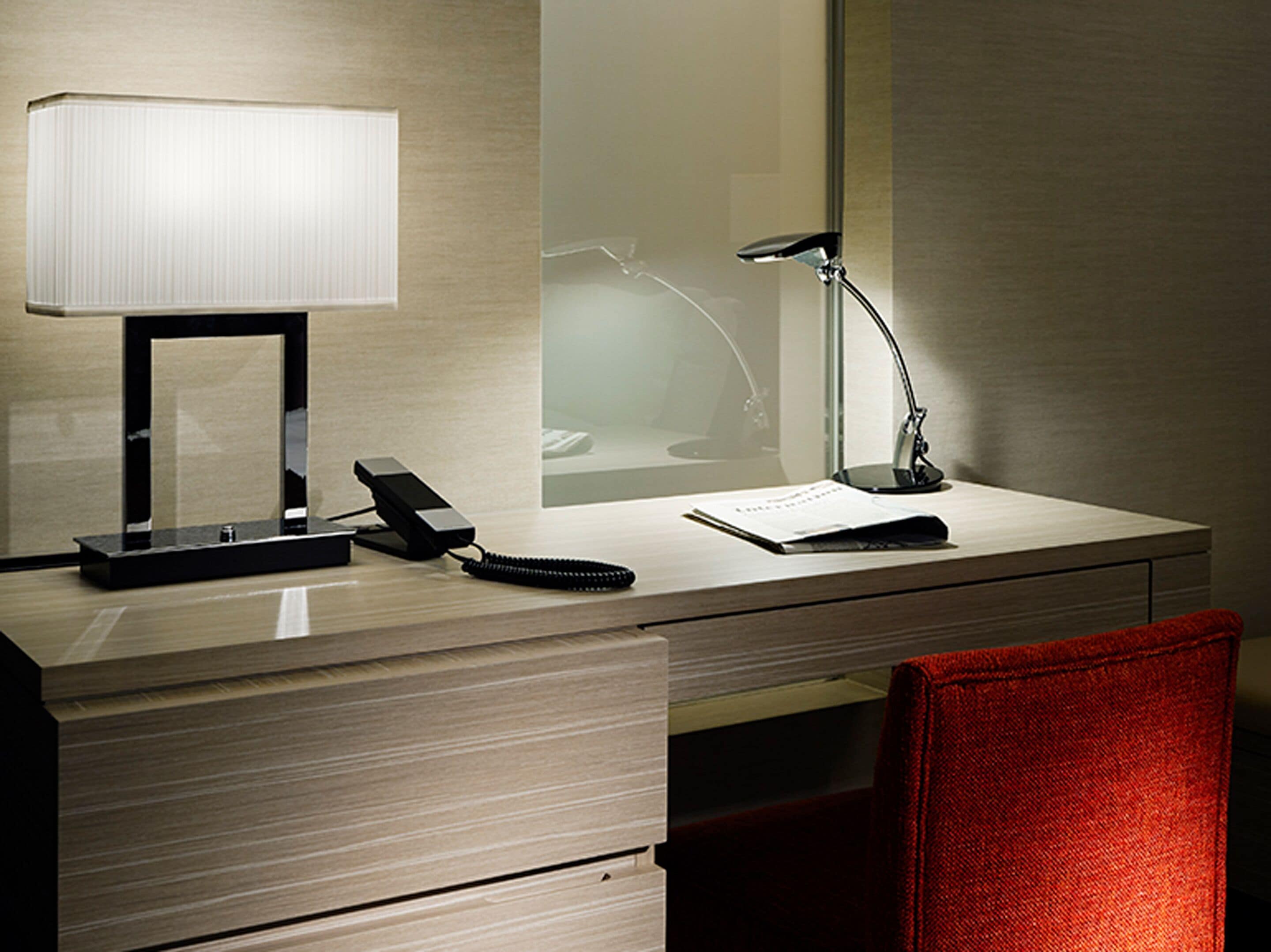 Et hotelværelse med et skrivebord, telefon, lampe, spejl og en rød stol.