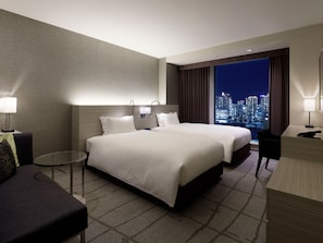 Quarto de hotel com duas camas, uma escrivaninha, uma cadeira, uma mesinha e vista para o horizonte urbano à noite.